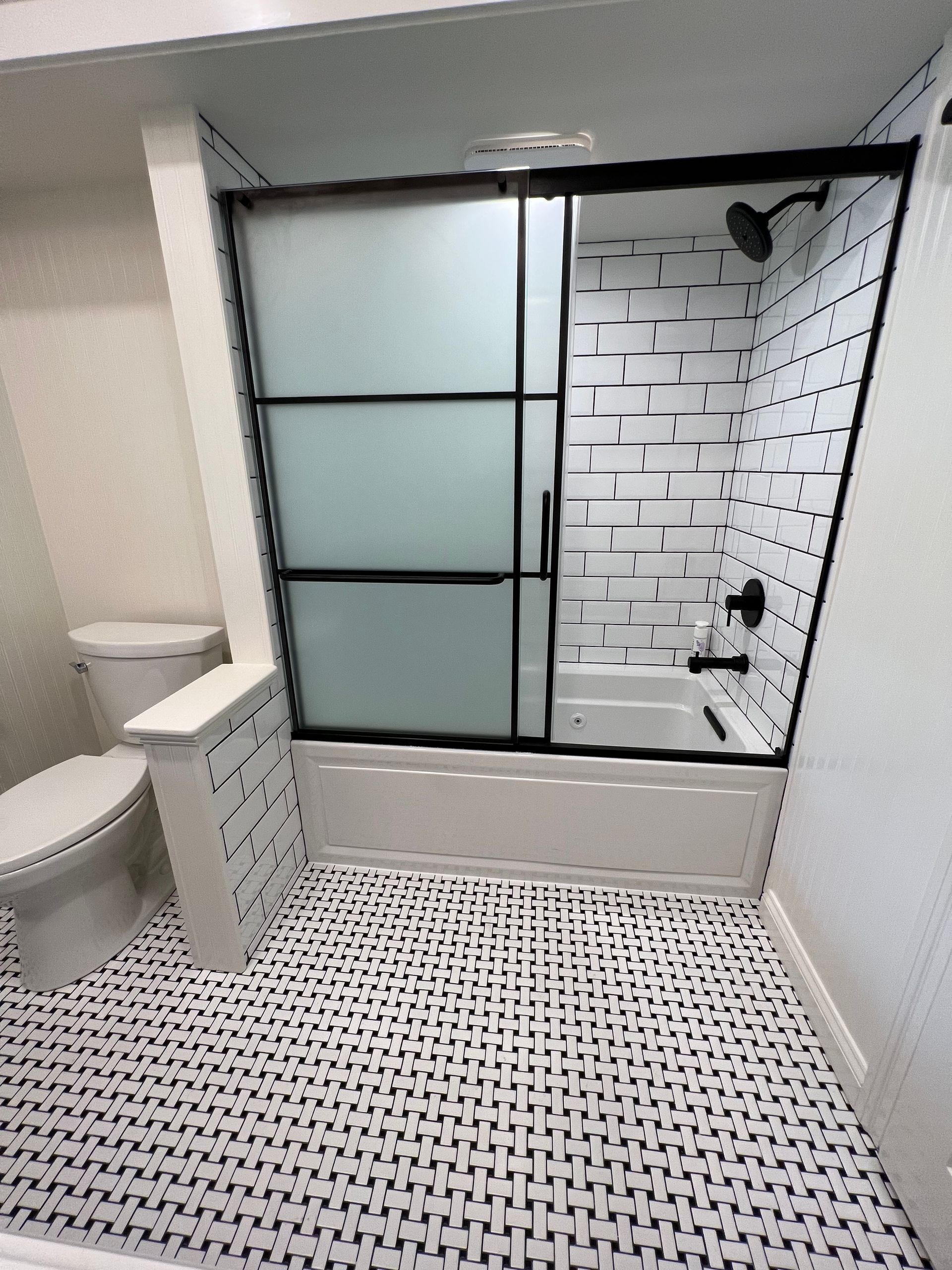 A bathroom featuring a patterned tiled floor, a white toilet, and a bathtub with black-framed glass sliding doors.