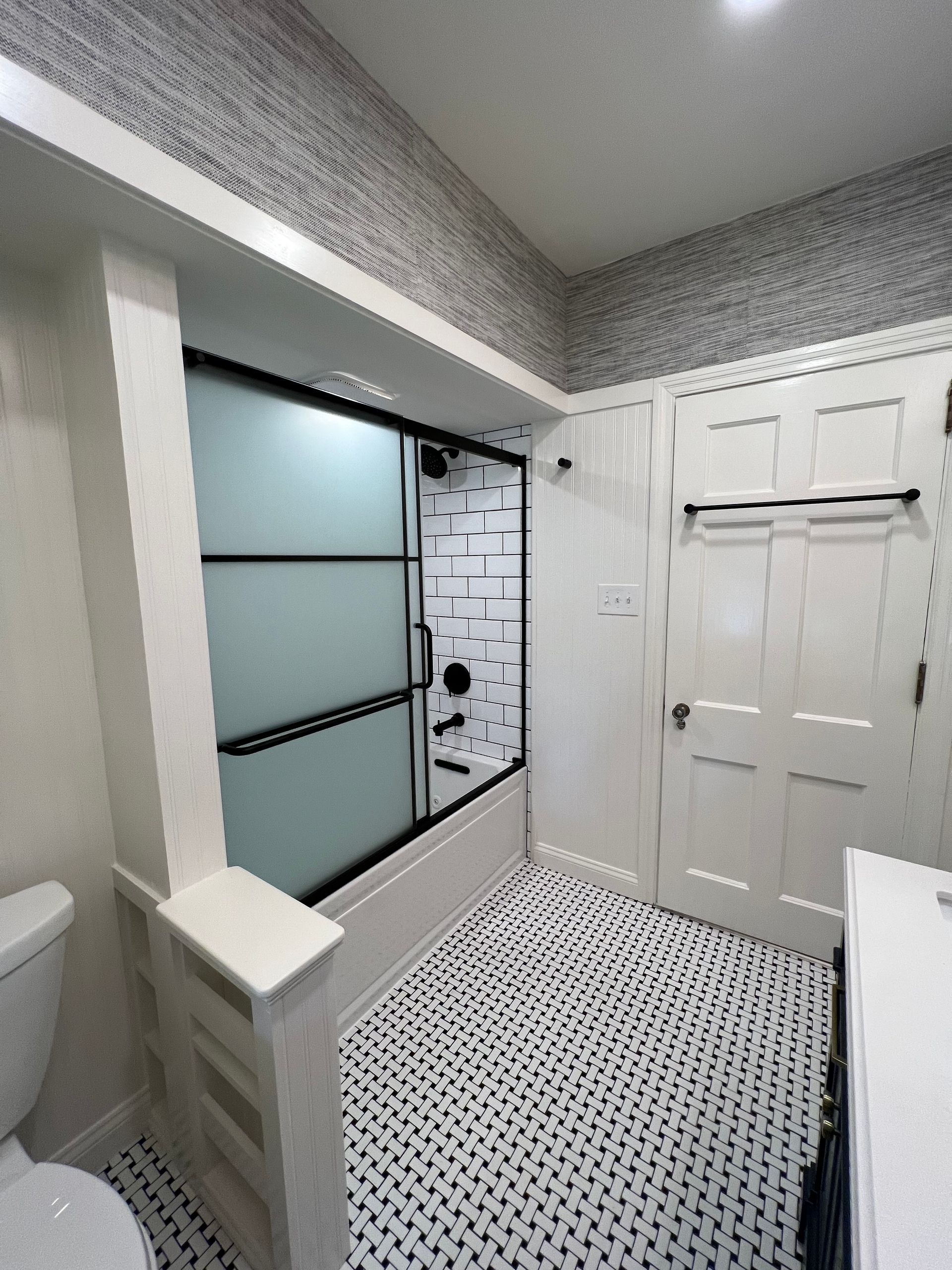 A bathroom with a shower enclosed by black-framed frosted glass, white wainscoting, and patterned black-and-white tile.