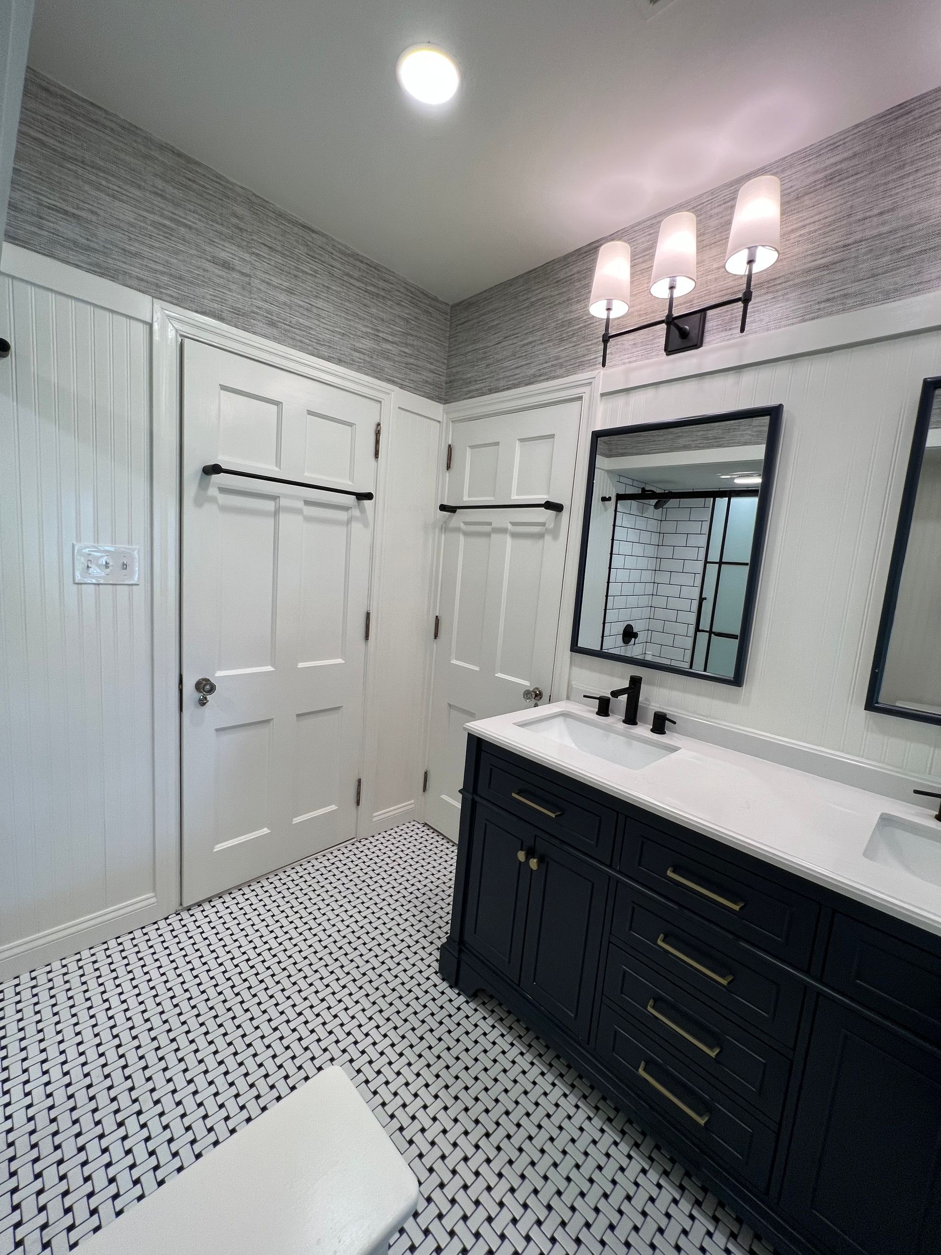 A modern bathroom features a dark blue vanity, a rectangular mirror, patterned floor tiles, and textured gray wallpaper.