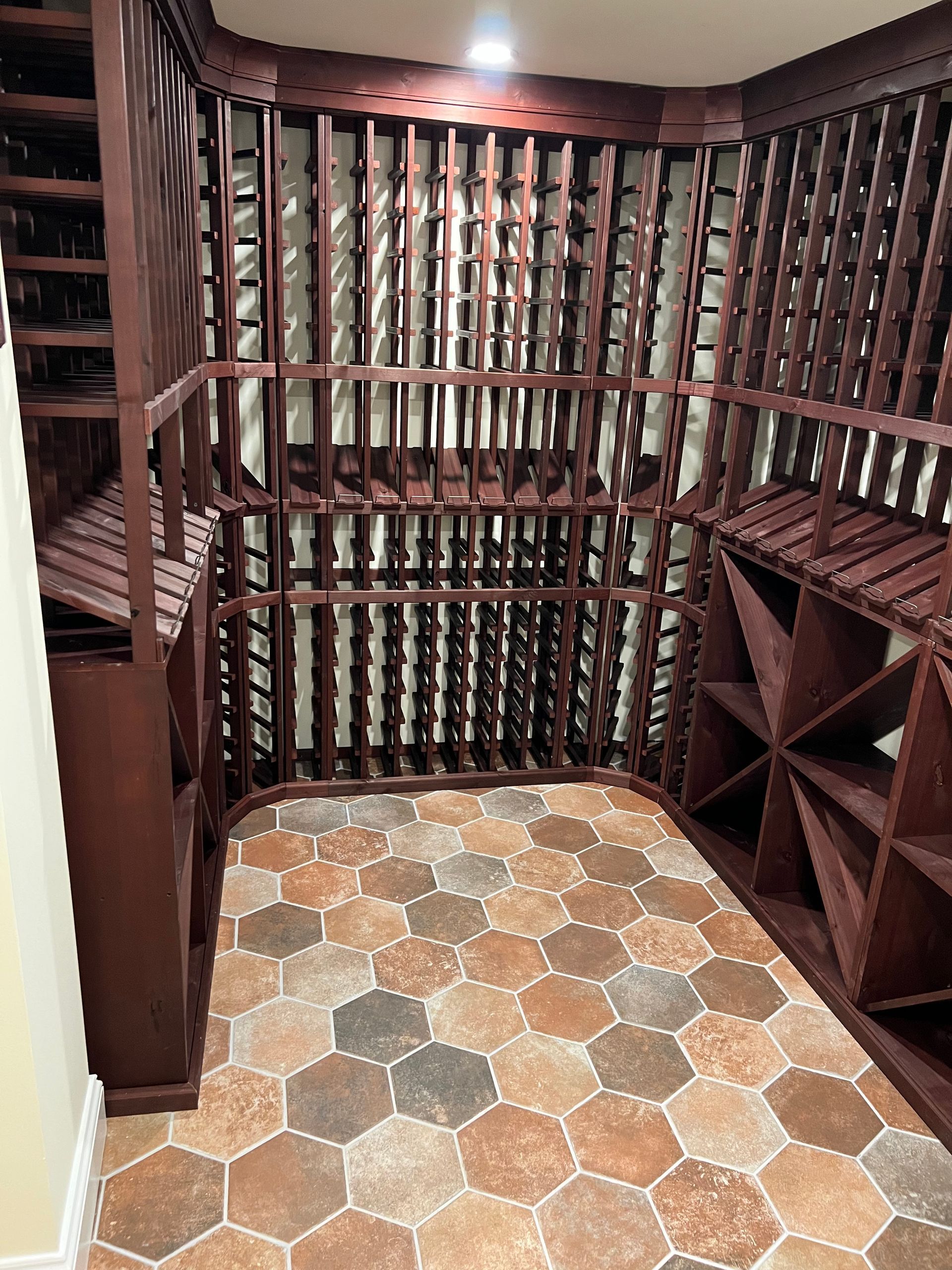 A small walk-in wine cellar with dark wood storage racks lining the walls and a brown hexagonal tiled floor.