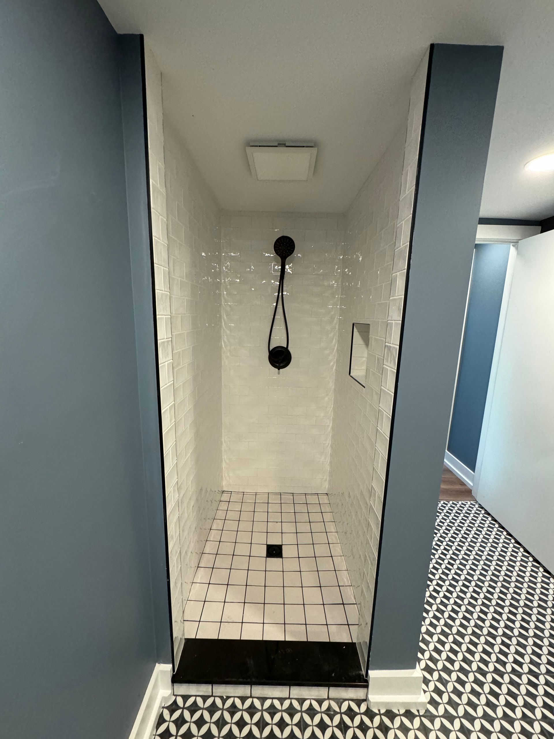 A walk-in shower with white tiled walls, a black handheld showerhead, and a black-and-white patterned floor.