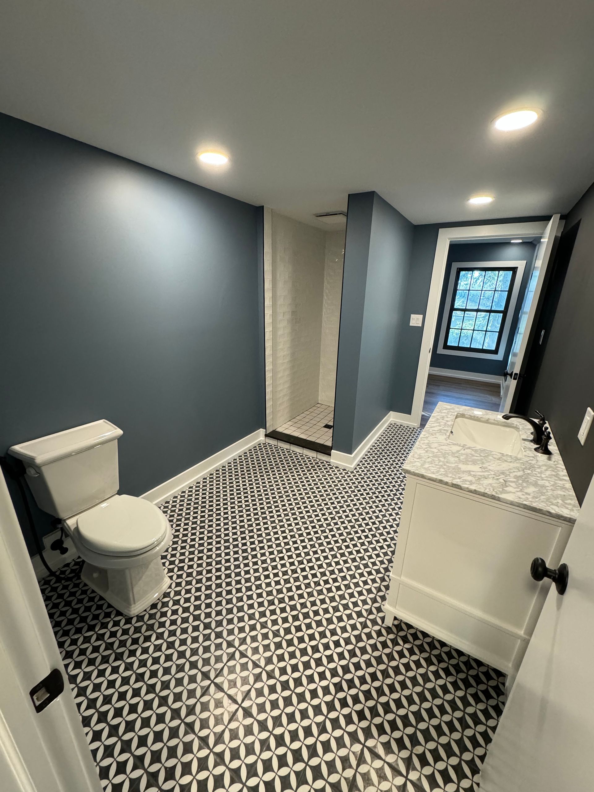 A modern bathroom with dark blue walls, a white vanity with marble top, a toilet, and patterned black-and-white tile.