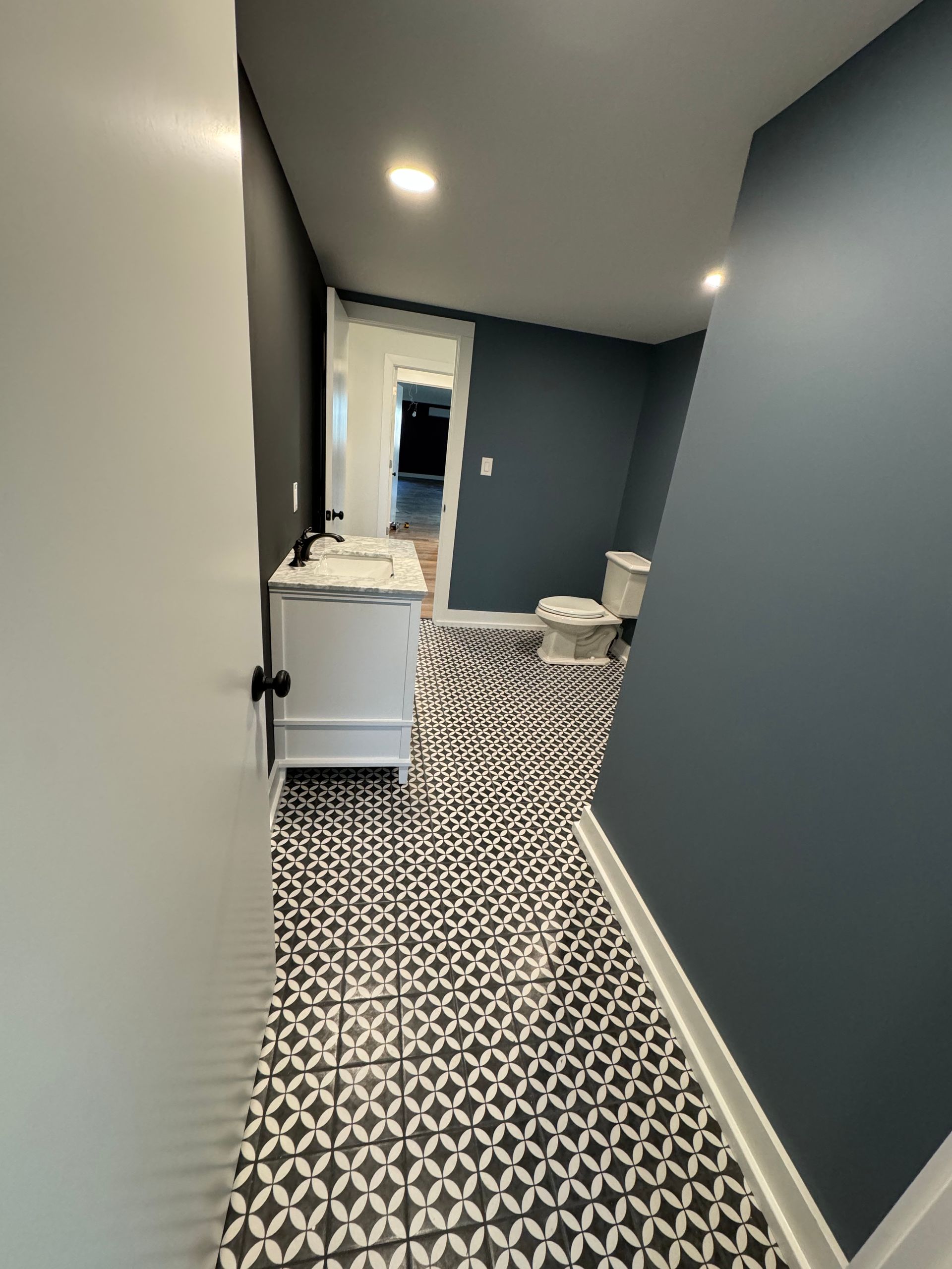 A bathroom with navy walls, patterned tile floors, a white vanity, and a toilet, seen through an open white door.