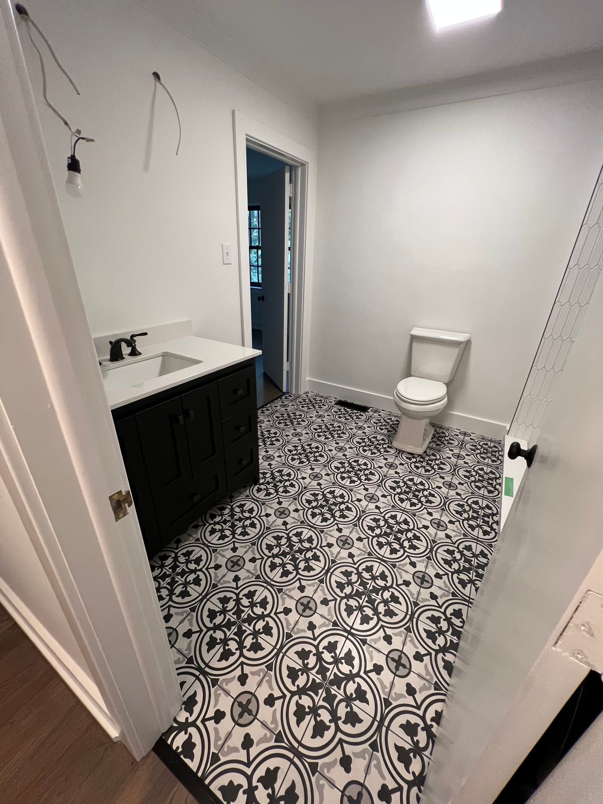 A bathroom with patterned black-and-white floor tiles, a black vanity, and a white toilet against a light wall.