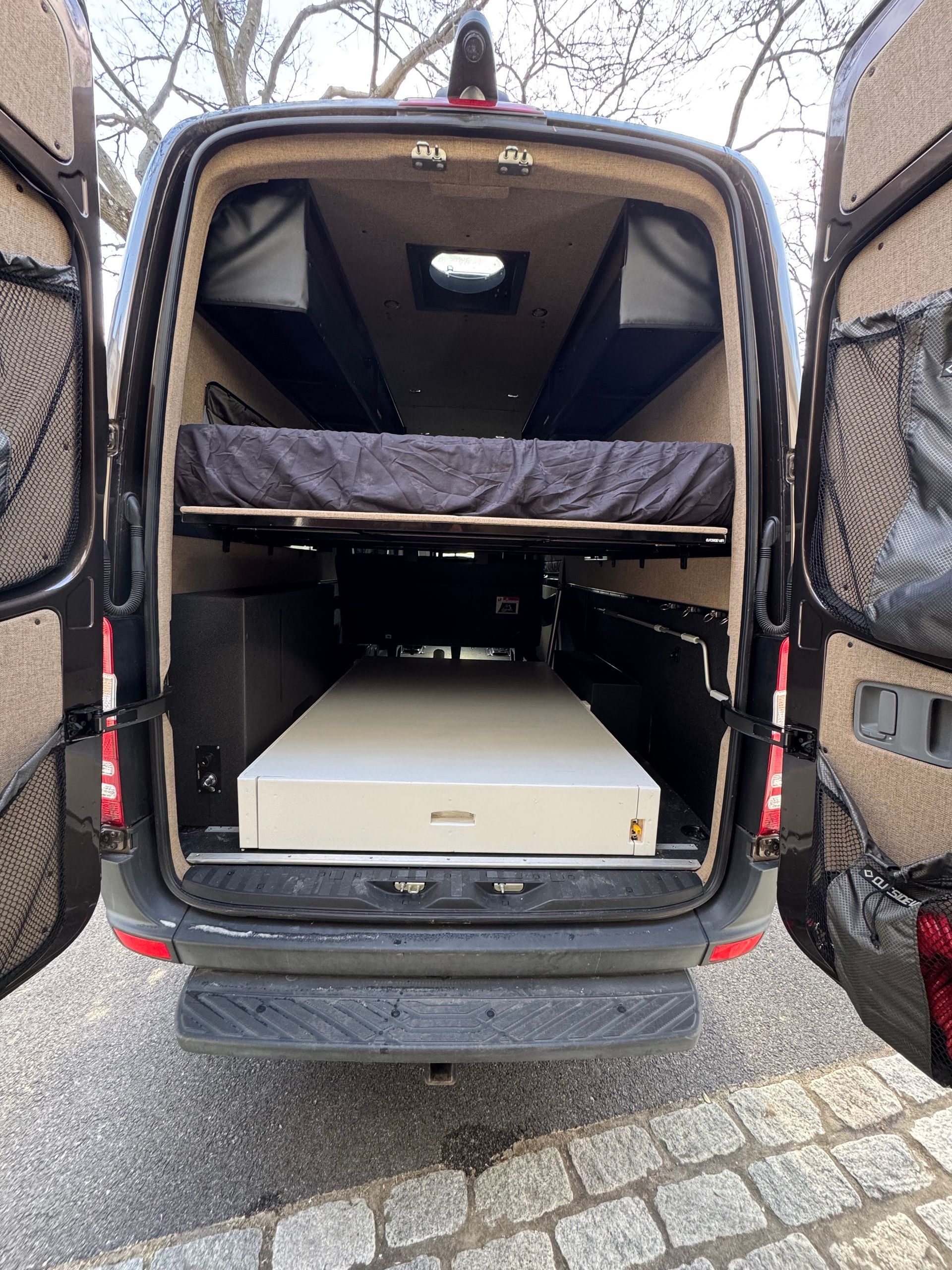 Open rear doors of a van showing a lower cargo area with a white drawer unit and an elevated platform bed with a mattress.