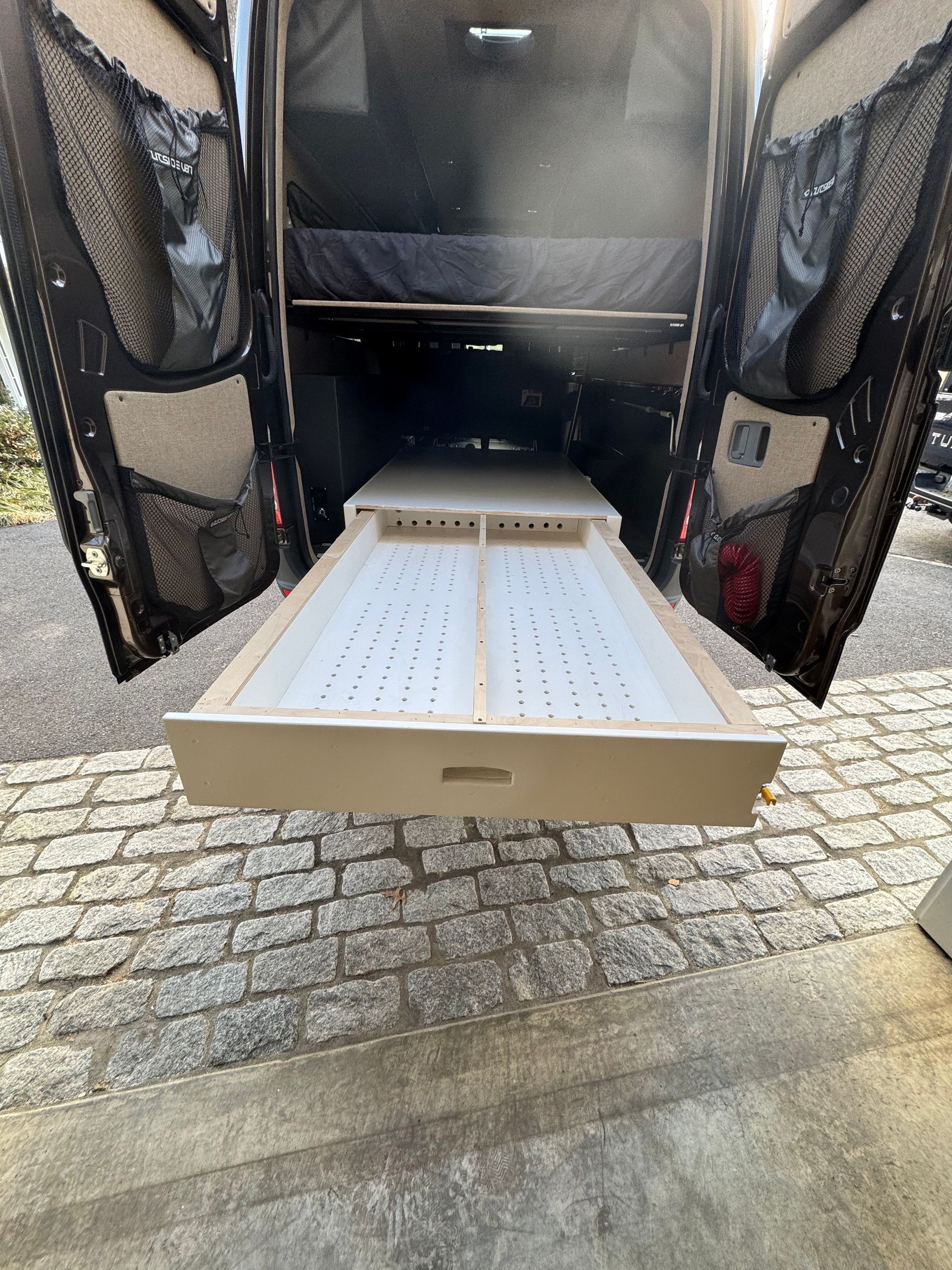 Rear view of a van interior featuring a large, beige pull-out storage drawer with a ventilated base extended outward.