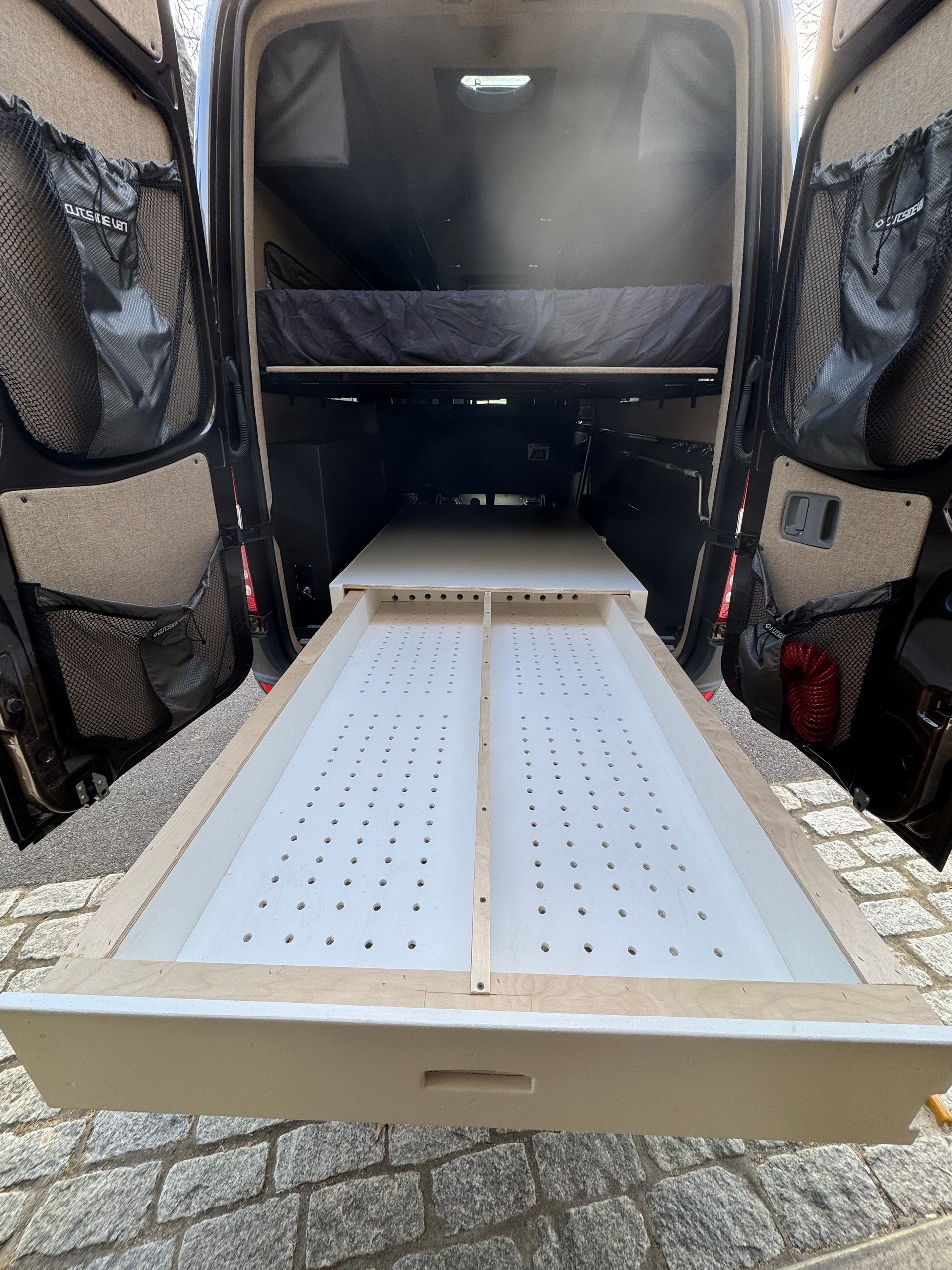 A pull-out drawer with a perforated white base installed in the rear storage area of a van under a raised bed platform.