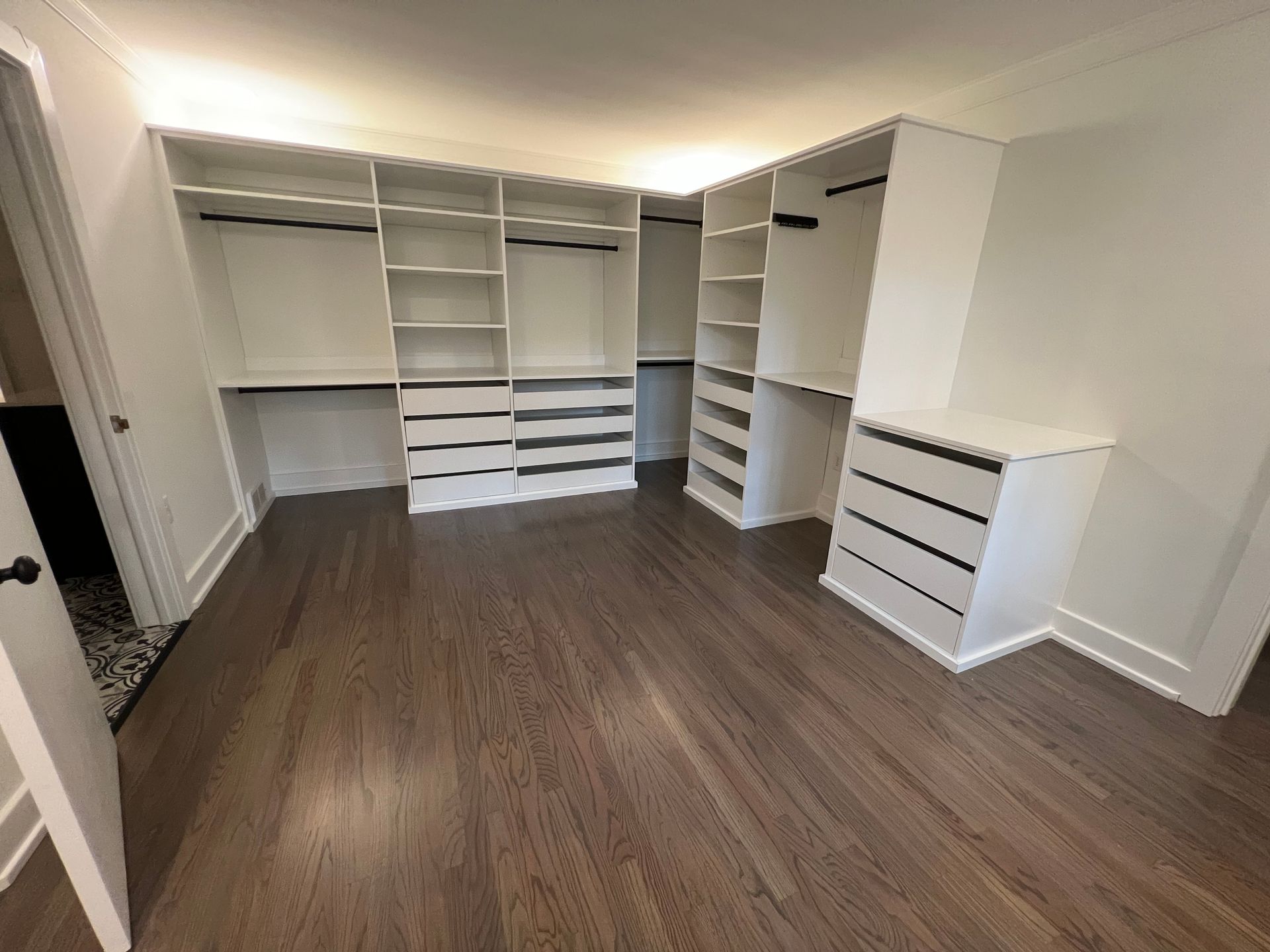 A white walk-in closet with built-in shelves, hanging rods, and drawers, set against a room with dark wood flooring.