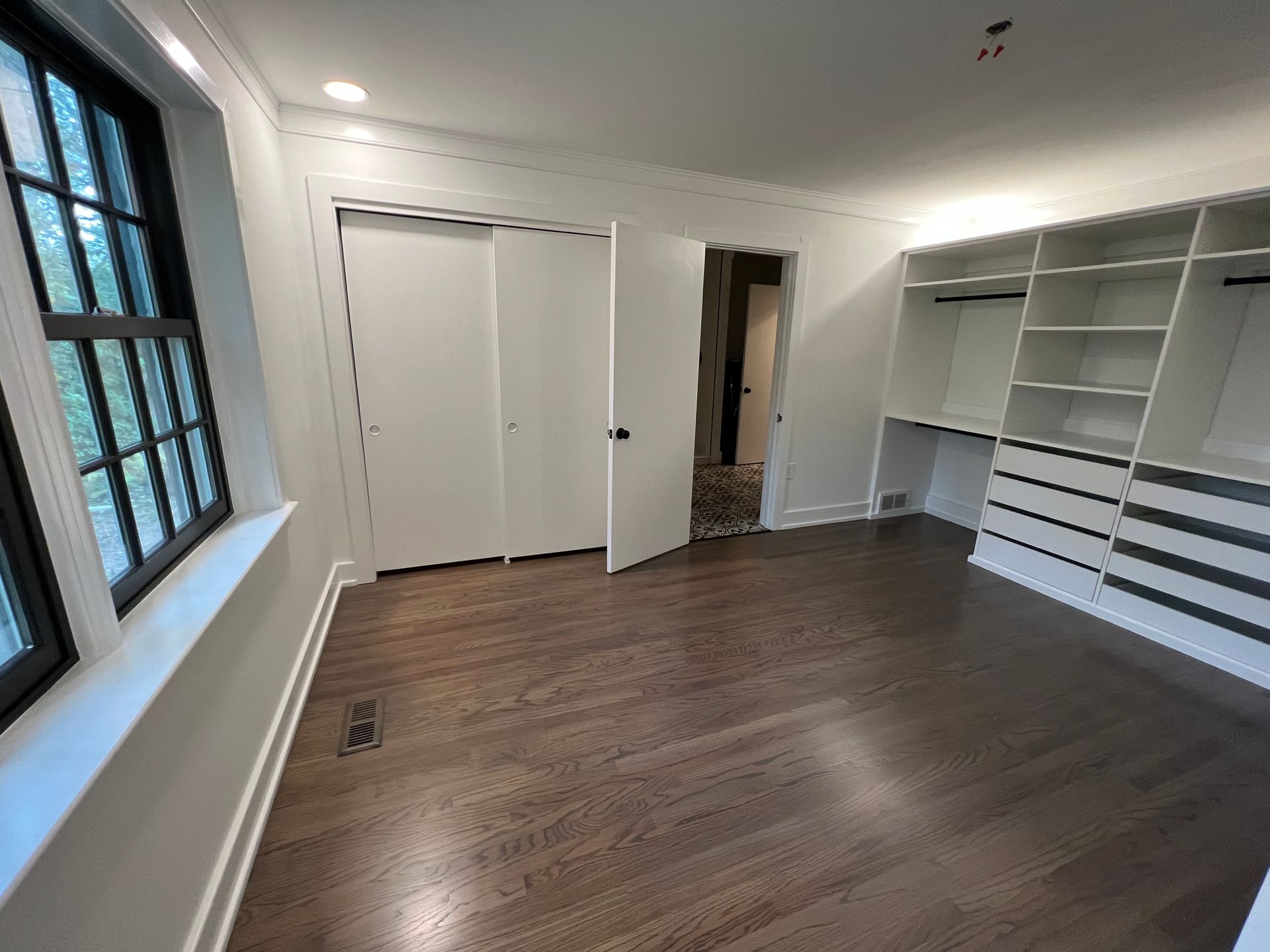 A room with dark wood flooring, a black-framed window, white closet doors, and built-in shelving with drawers.