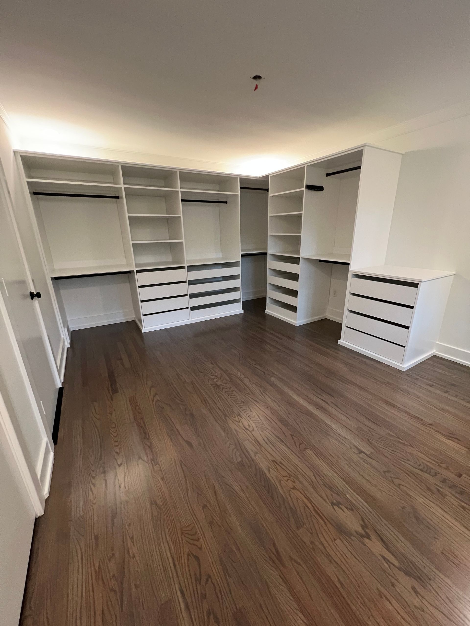 A walk-in closet featuring white, custom-built storage units with shelving, hanging rods, and drawers over dark wood floors.