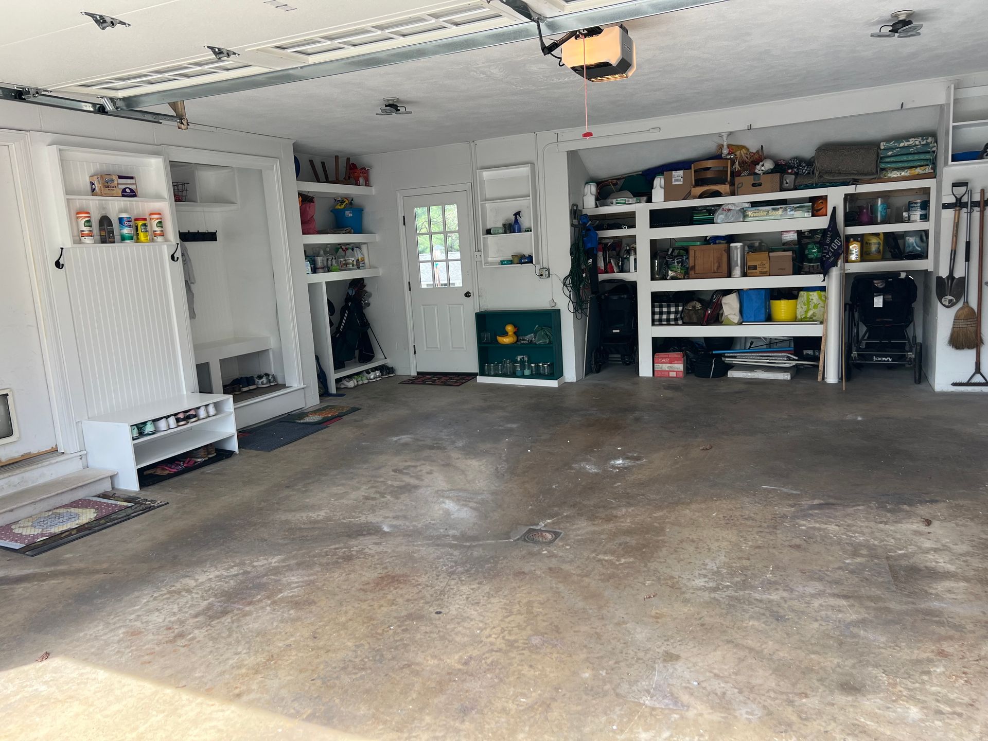 A white, cluttered garage with storage shelving units on the walls, a white door, and a stained concrete floor.