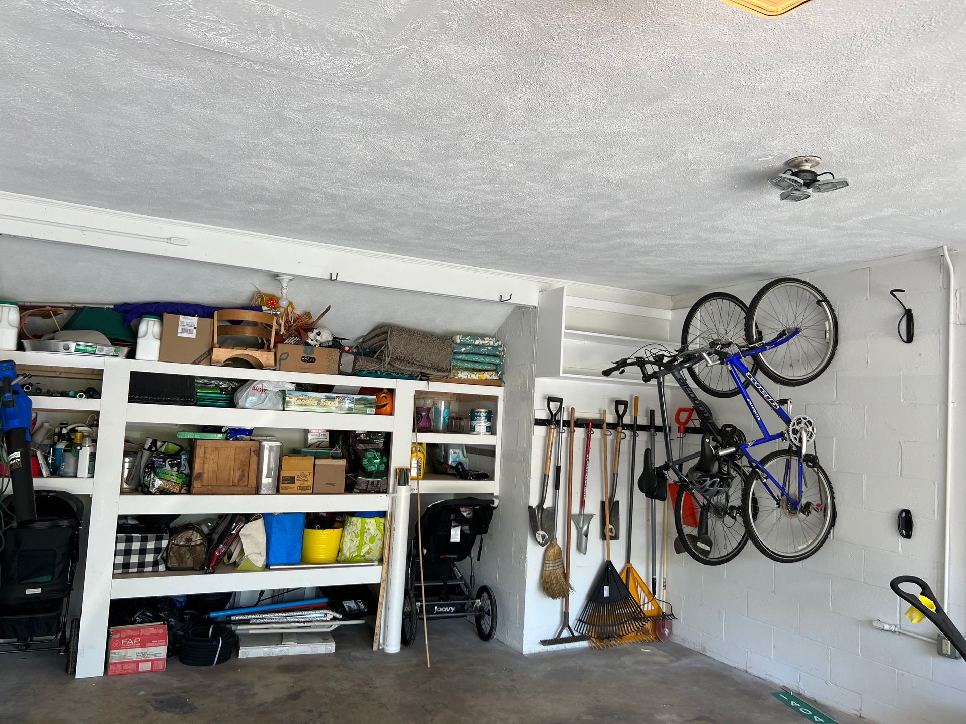 A garage interior showing storage shelves filled with boxes, garden tools hanging on a rack, and a bike mounted on the wall.