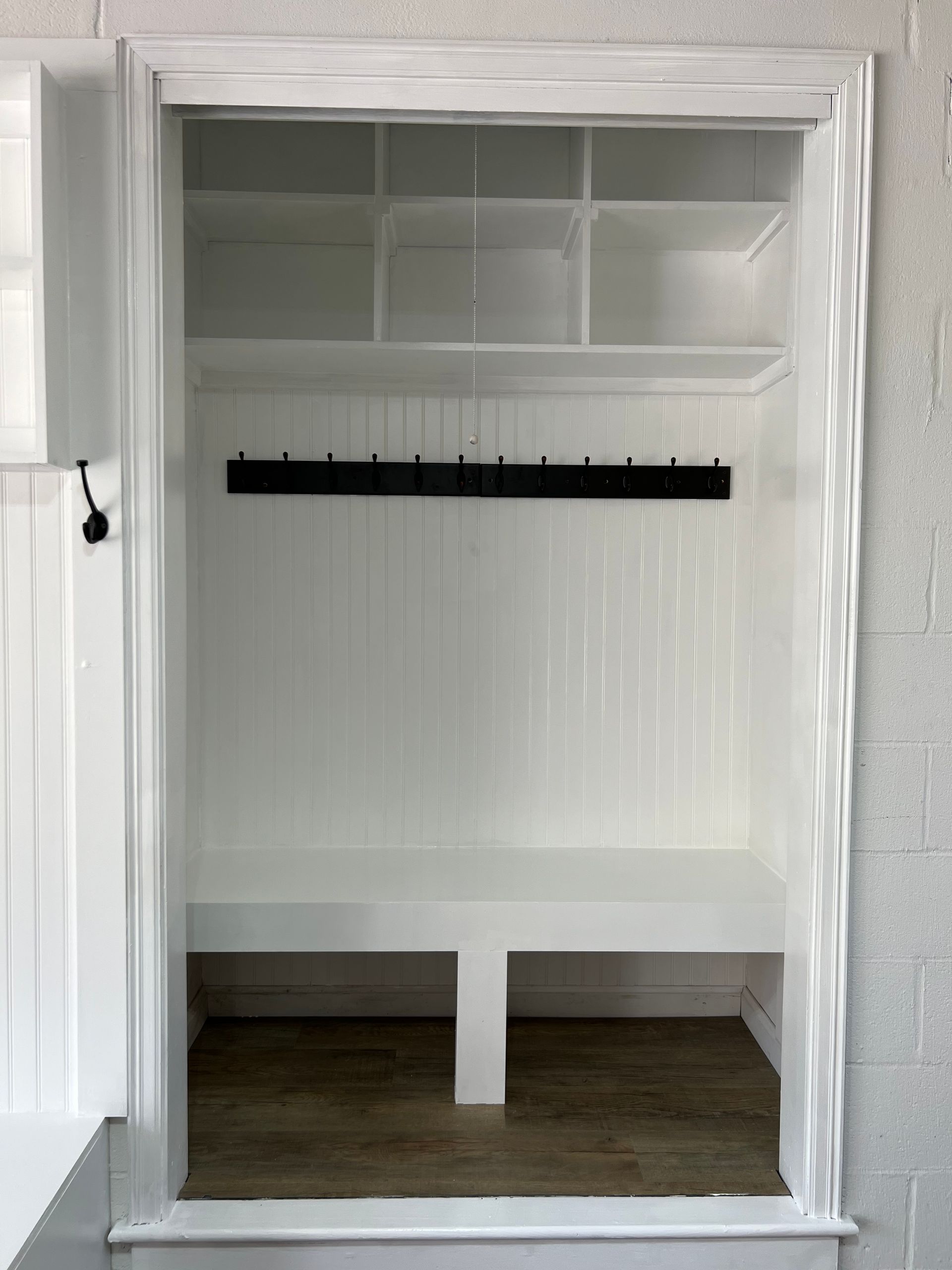 A white, built-in mudroom nook with cubbies, a black row of coat hooks, a bench seat, and wood-look flooring.