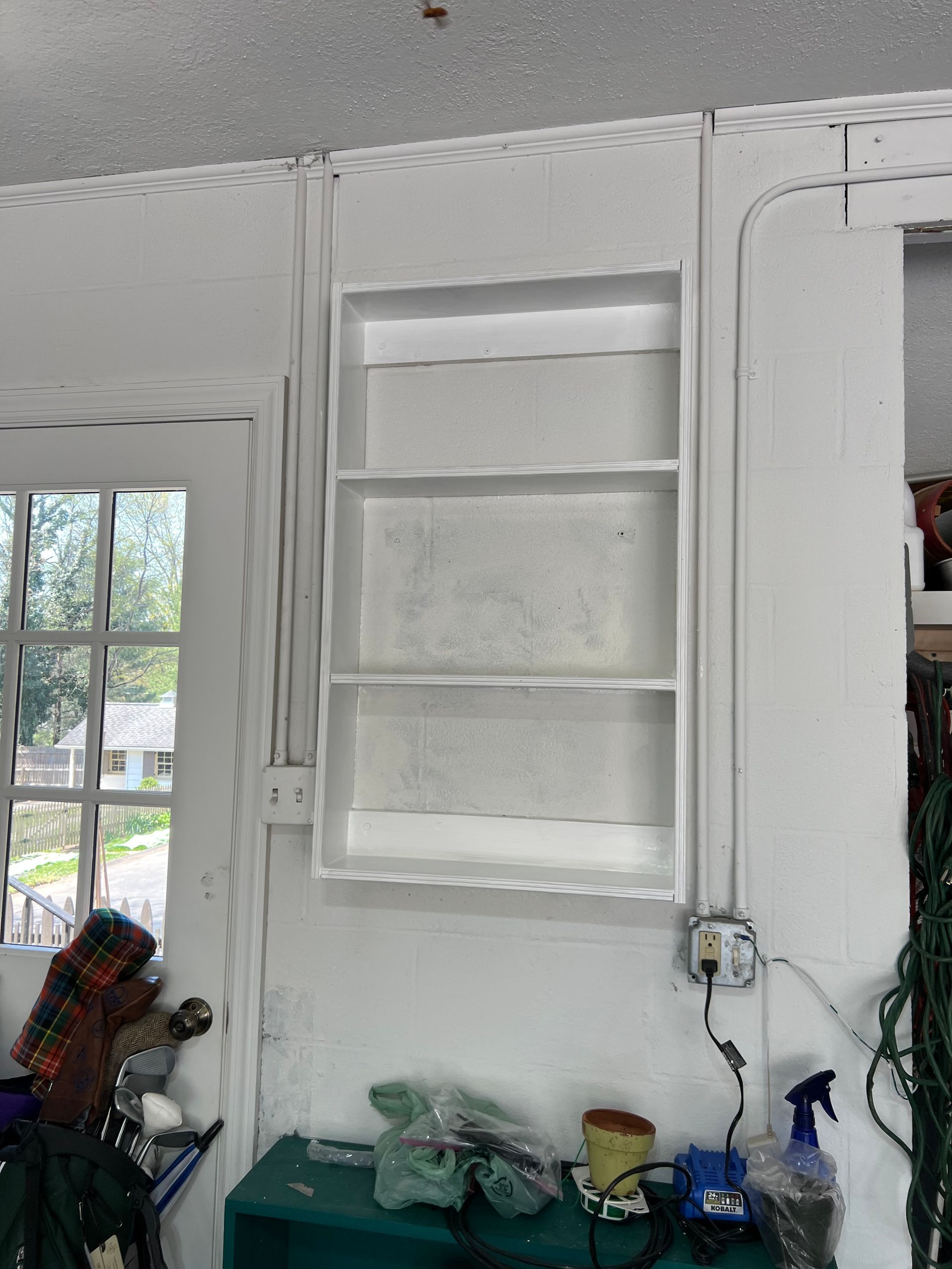 A white, three-tiered wall shelf mounted next to a glass door in a garage with exposed electrical conduit and outlets.