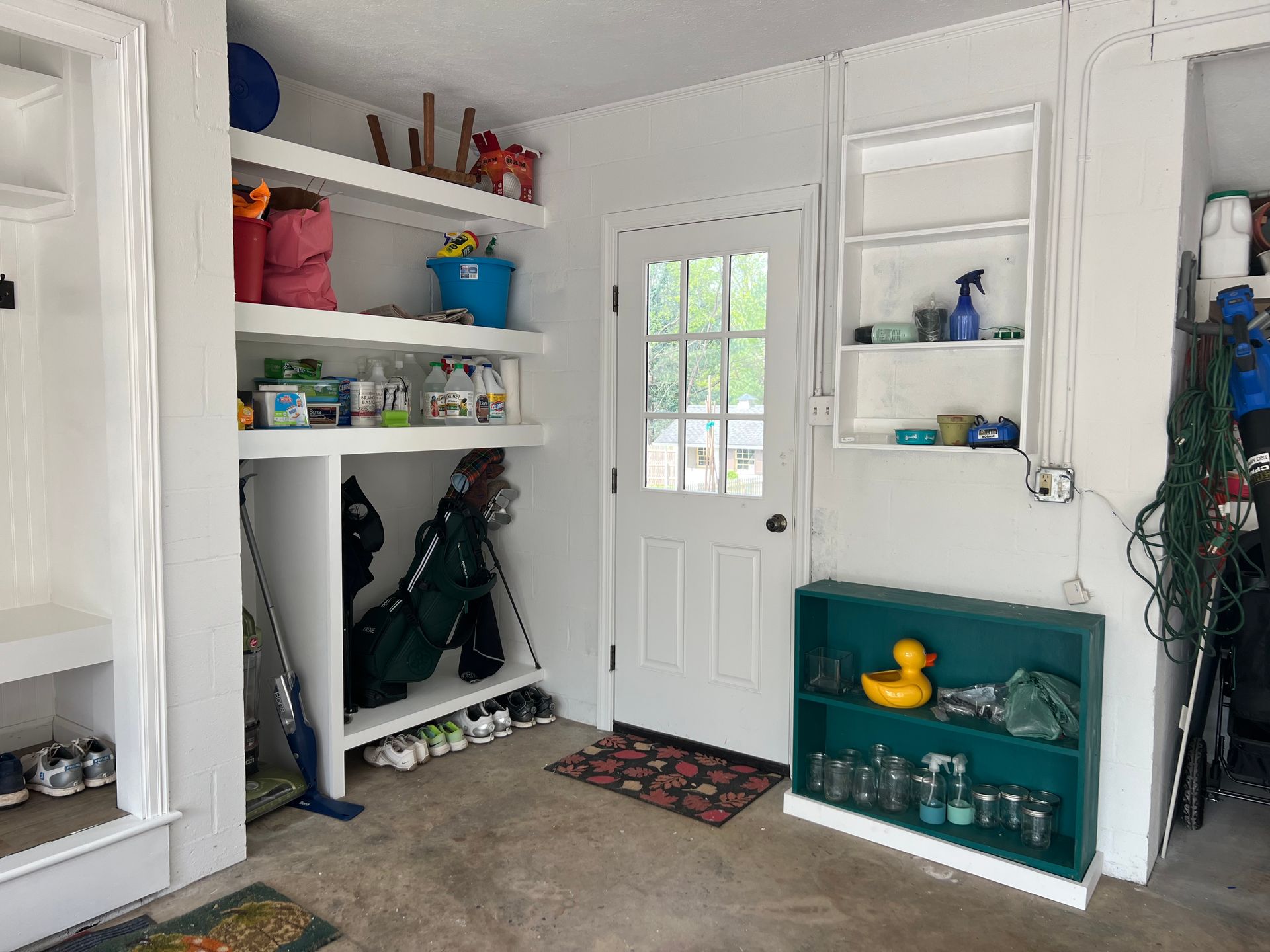 Organized white shelves line the walls of a garage, featuring a white entry door, storage bins, and a teal shelving unit.