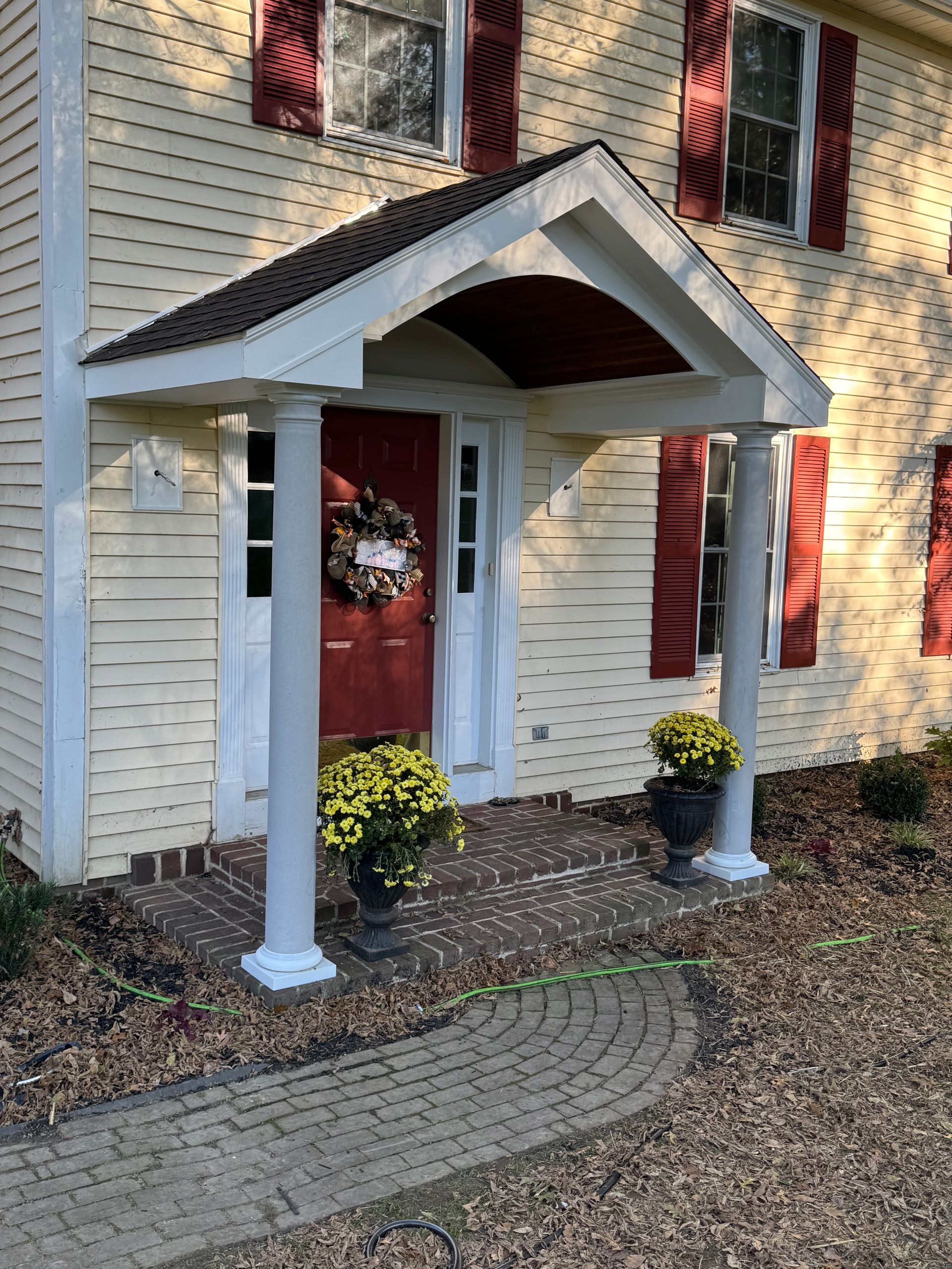 Yellow house exterior featuring a front porch with white columns, a red door with a wreath, and two pots of yellow mums.