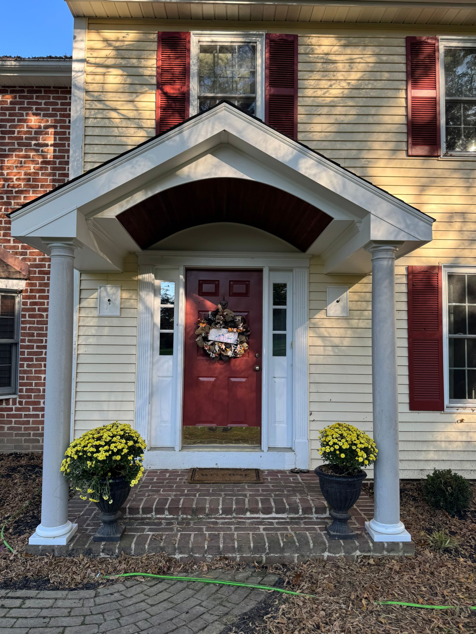 A yellow house with a covered entryway, red door, brick walkway, and potted yellow mums on the front porch.