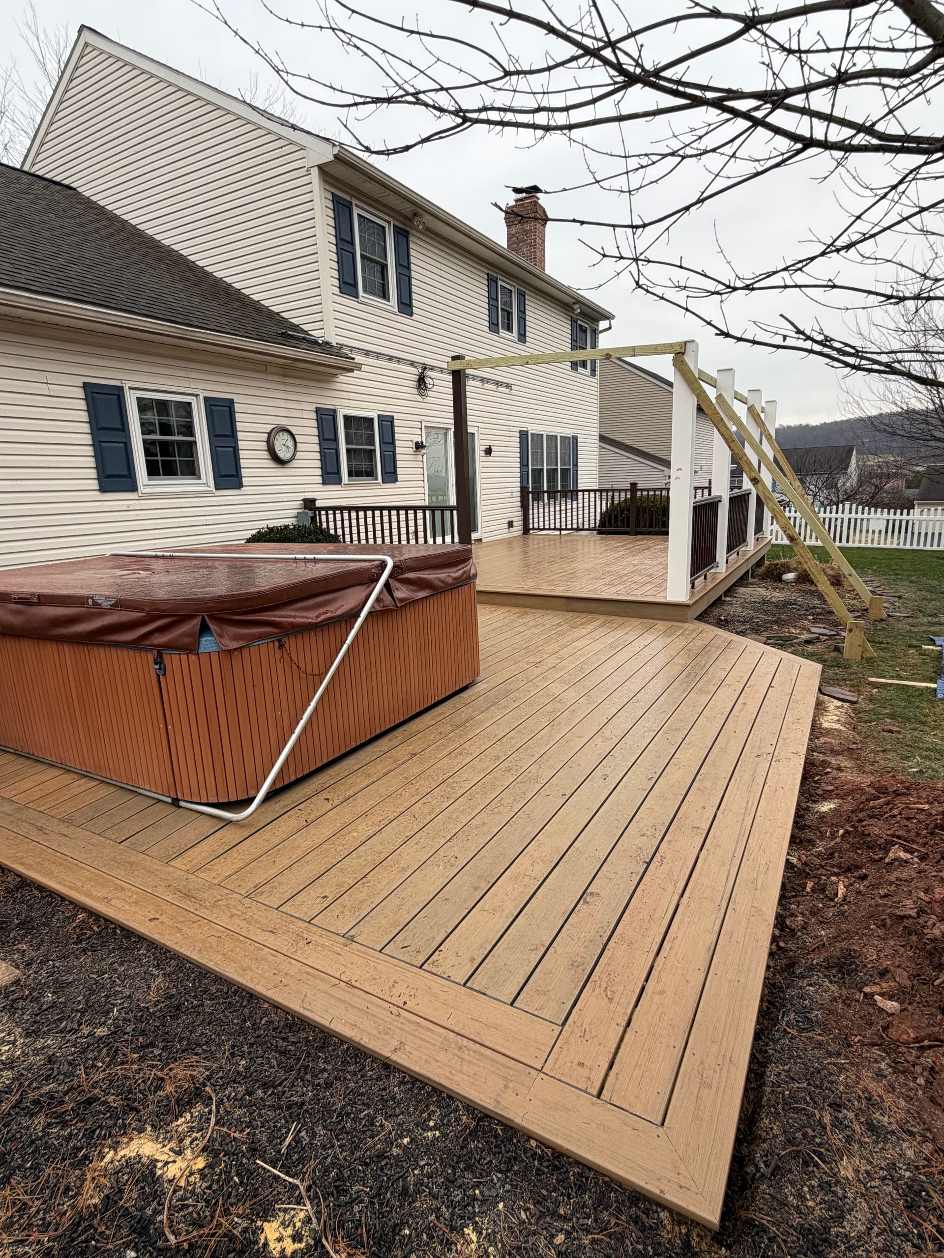 A tan deck featuring a hot tub with a brown cover sits beside a multi-story house with beige siding and blue shutters.