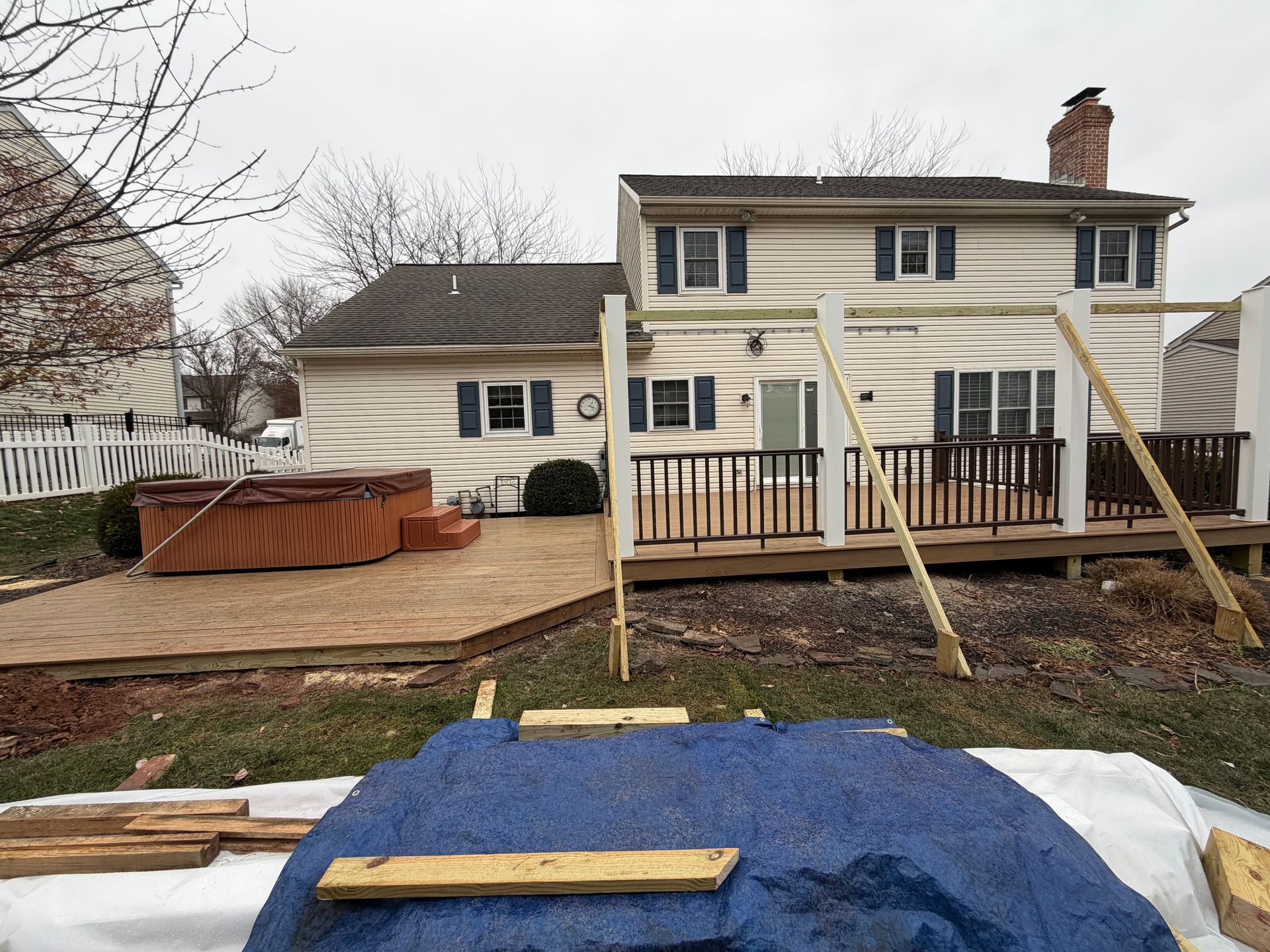 A two-story house with a wooden deck, a hot tub, and an outdoor pergola structure under construction in the backyard.
