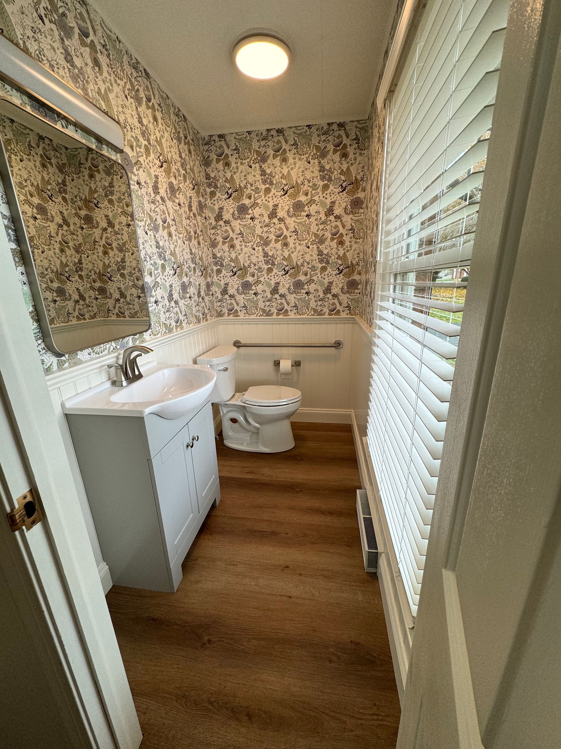 A small bathroom with floral wallpaper, a grey vanity with a white sink, a toilet, and a window with white blinds.
