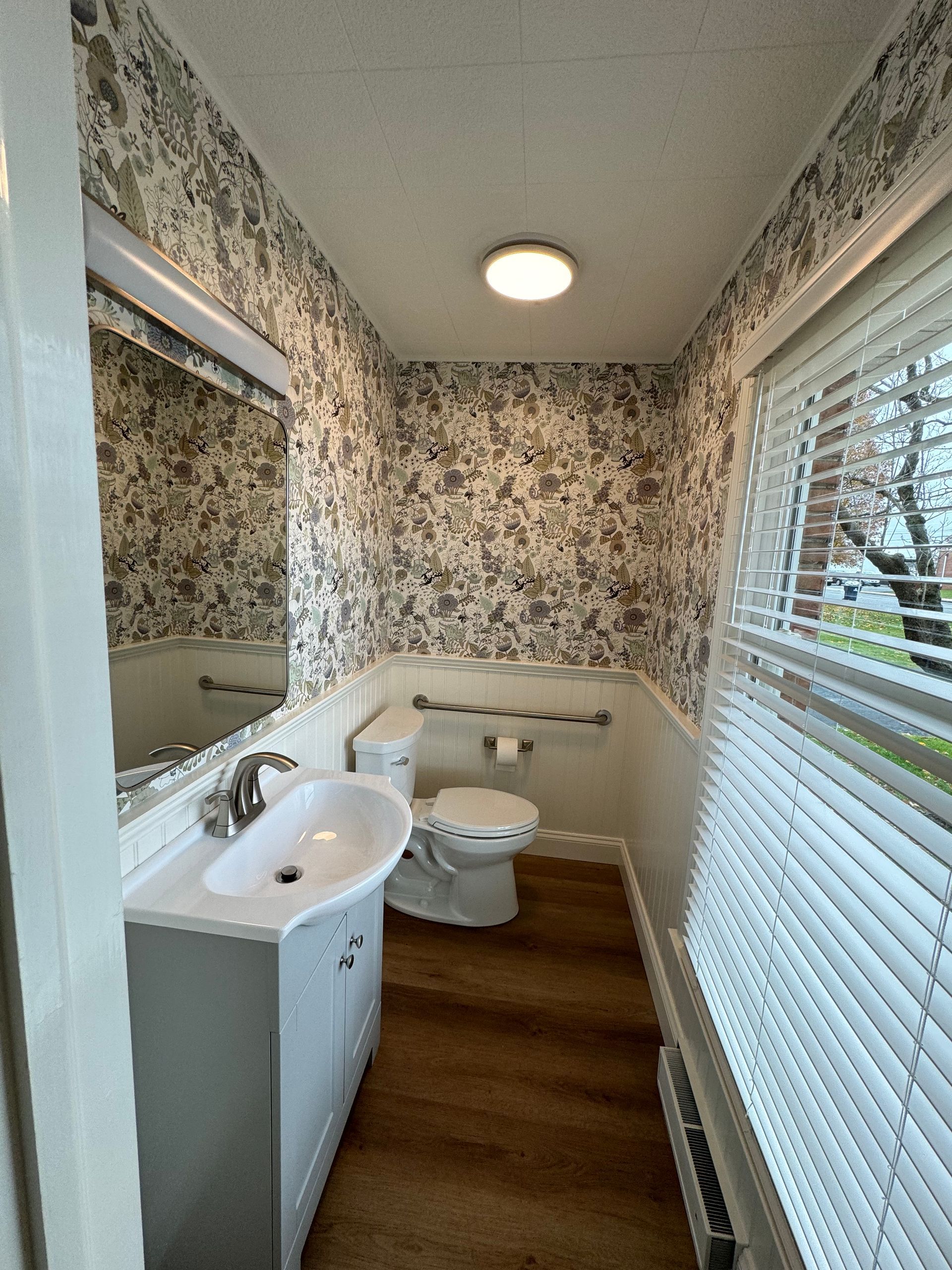 A small bathroom with floral wallpaper, a grey vanity with a white sink, a toilet, and a window with white blinds.