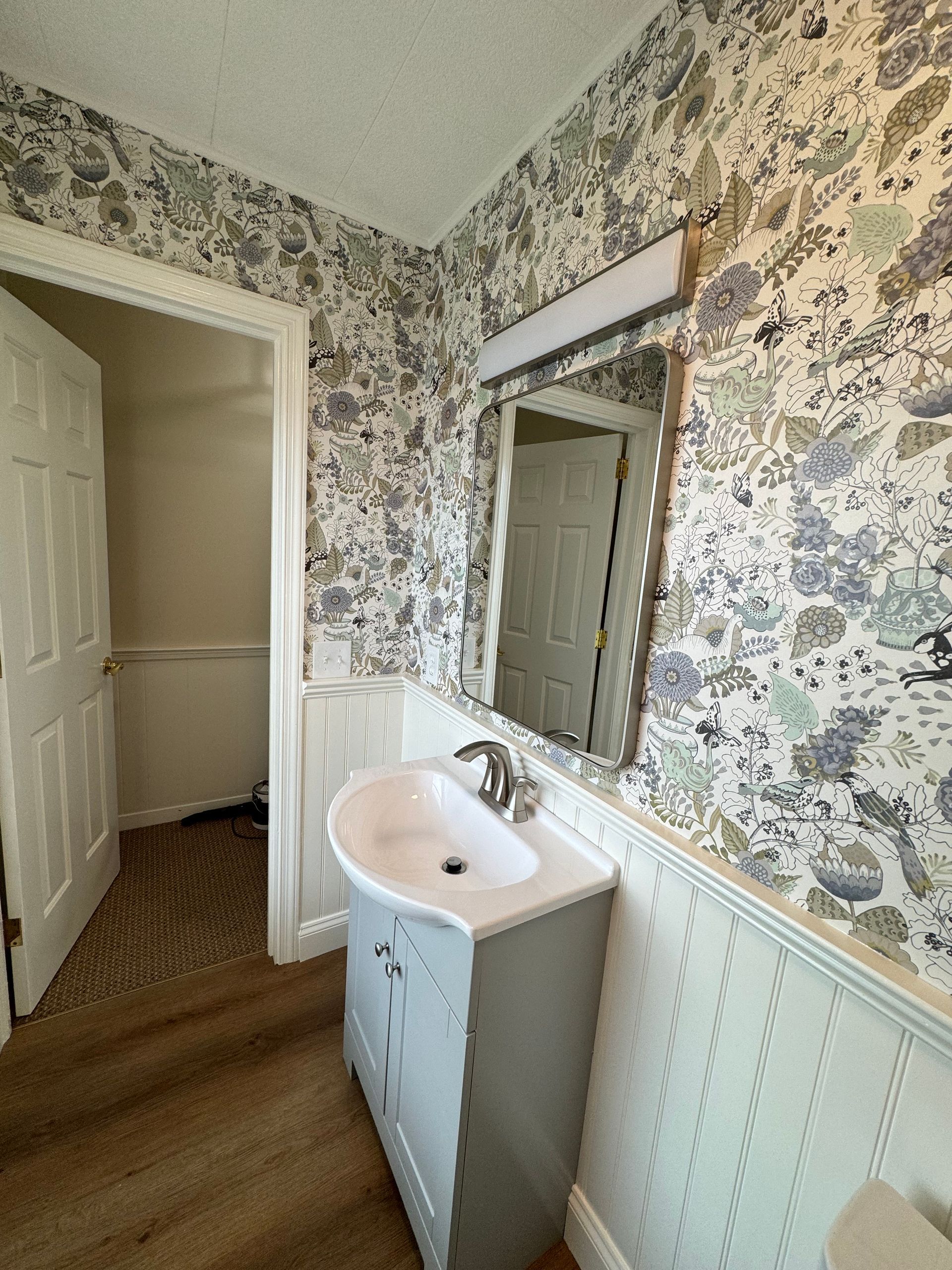 A bathroom vanity with a white basin and grey cabinet beneath a mirror, set against floral wallpaper and white wainscoting.