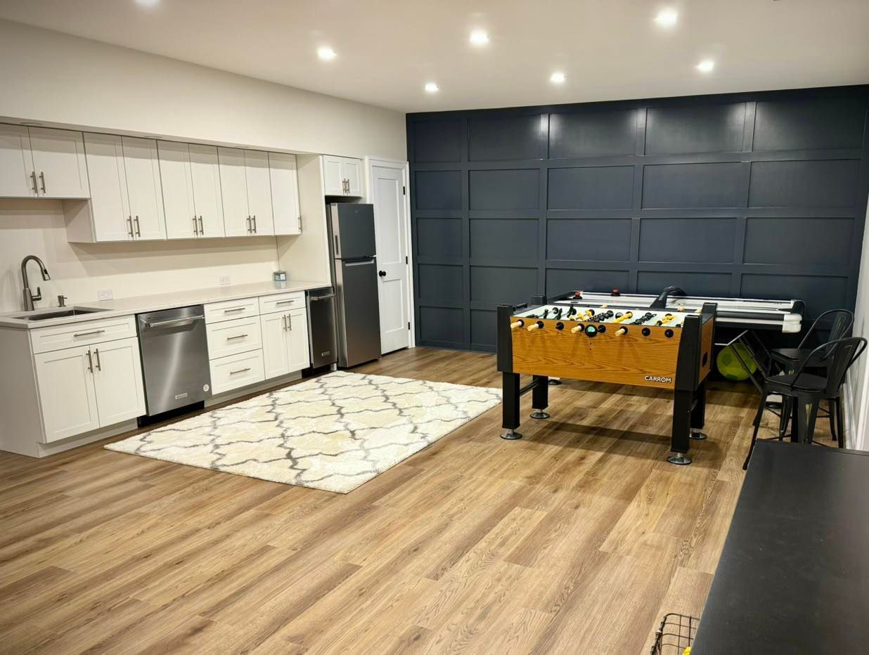 A finished basement with a kitchen area, a white patterned rug, and a foosball table against a dark blue paneled wall.