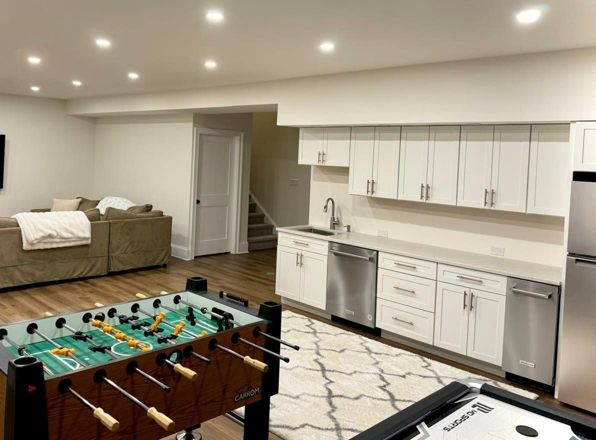 A basement with a foosball table, air hockey game, kitchen area with white cabinets, and a couch in the background.