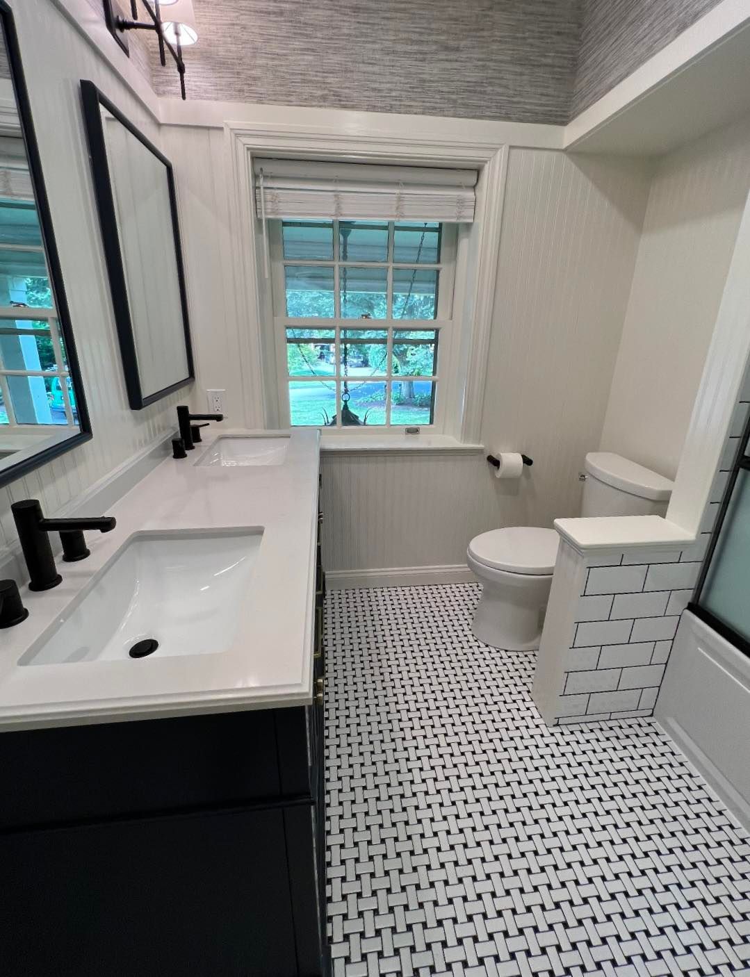 A modern bathroom with a double vanity, black fixtures, patterned tile floors, and a toilet beside a window.