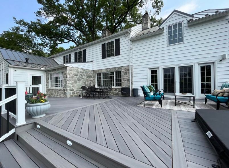A spacious gray composite deck with stairs leads to the back of a large white house with stone accents and patio furniture.
