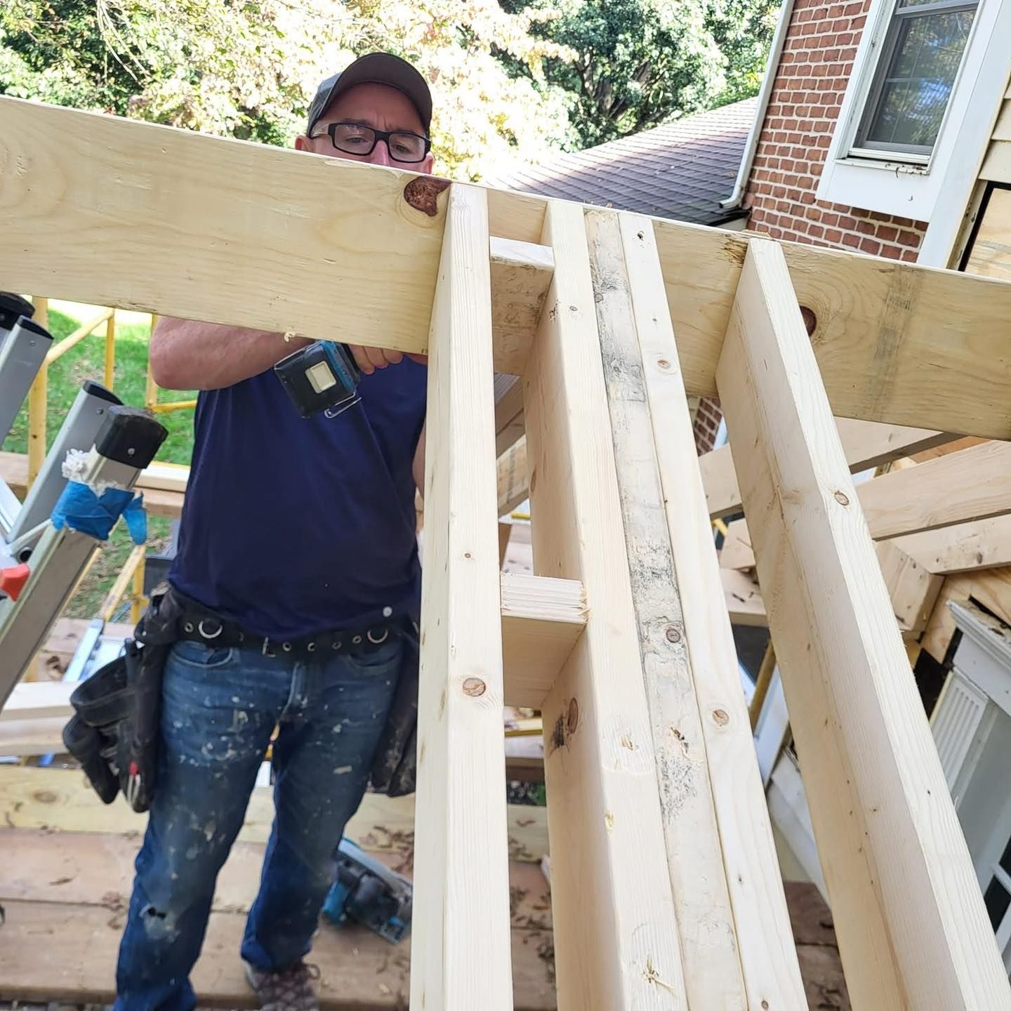 A person wearing glasses and a tool belt holds a large wooden beam during a construction project at a residential house.