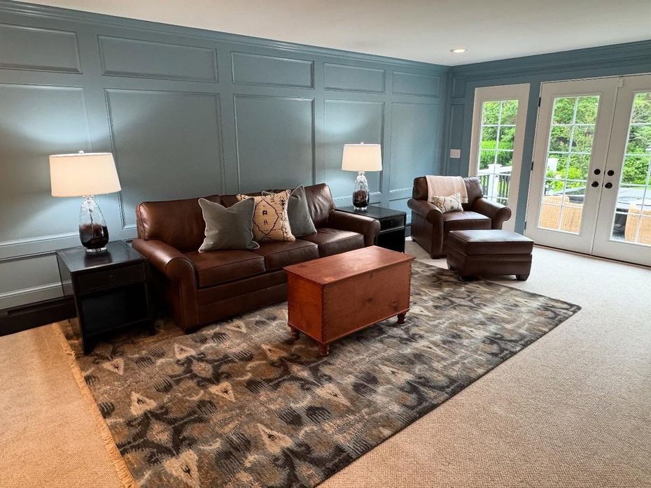 A living room with blue paneled walls, a brown leather sofa, matching armchair with ottoman, and a patterned area rug.