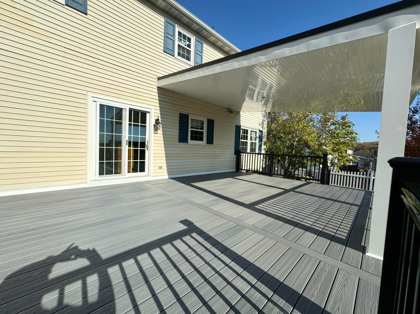 A gray composite deck with a white covered patio roof attached to a light yellow vinyl-sided house.