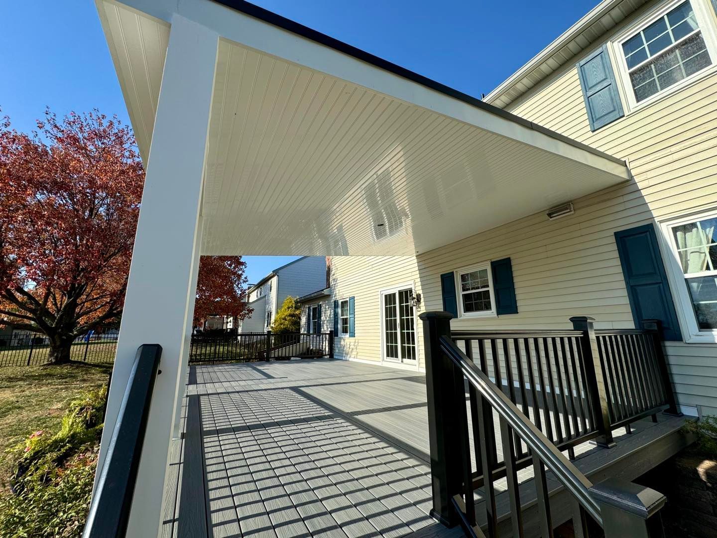 A covered backyard deck with light-colored pavers, a white ceiling, and dark railings against a beige house.
