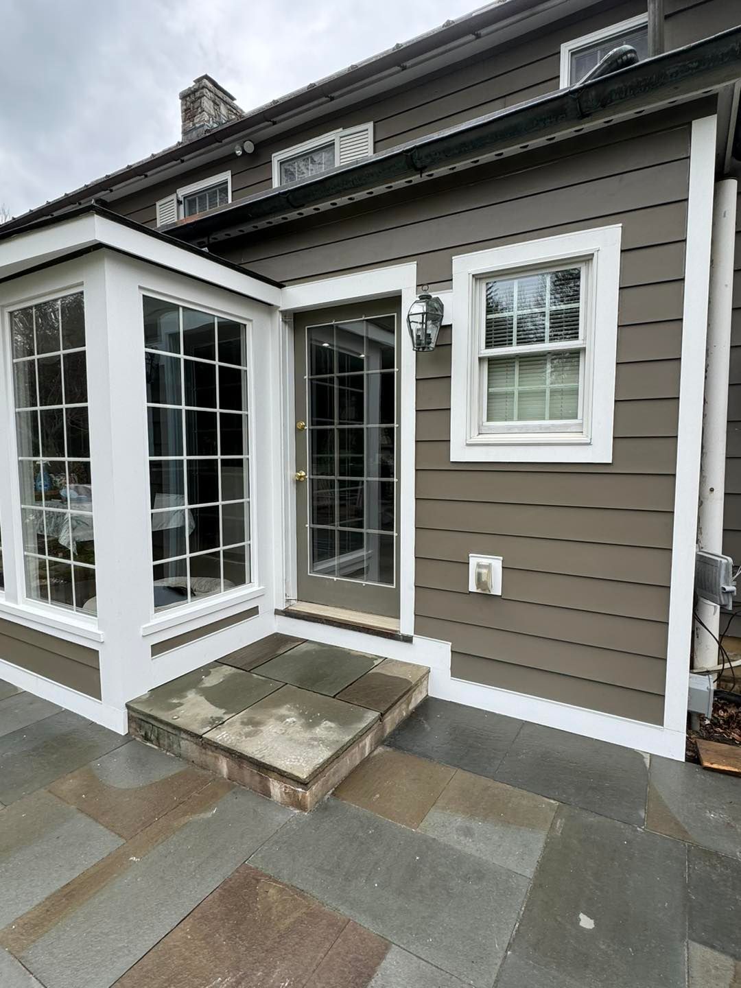 A brown house exterior featuring a small stone patio, a glass door, a bay window, and white trim against a cloudy sky.