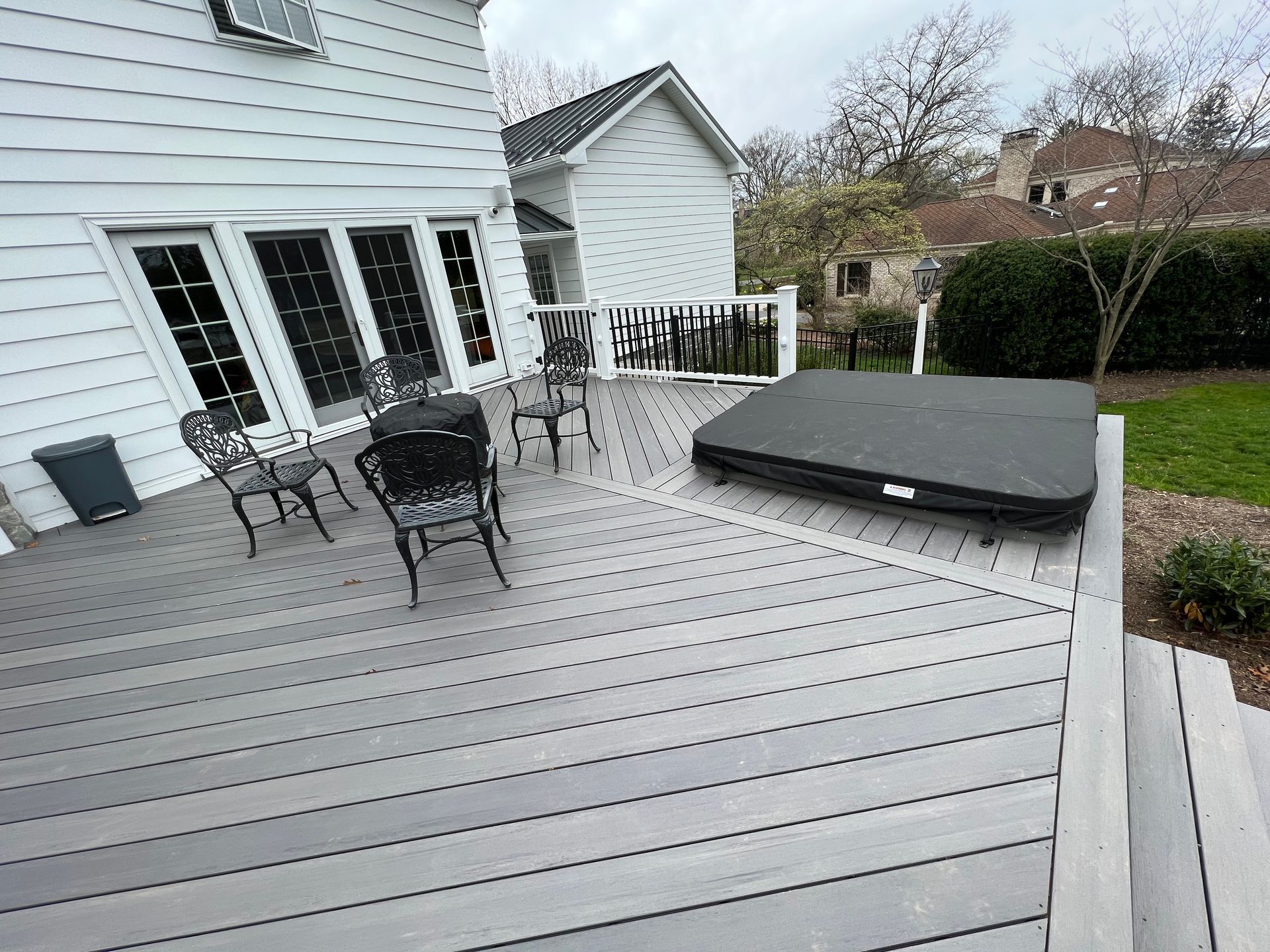 A custom composite deck featuring a hot tub with a black cover and a small seating area outside a white house.
