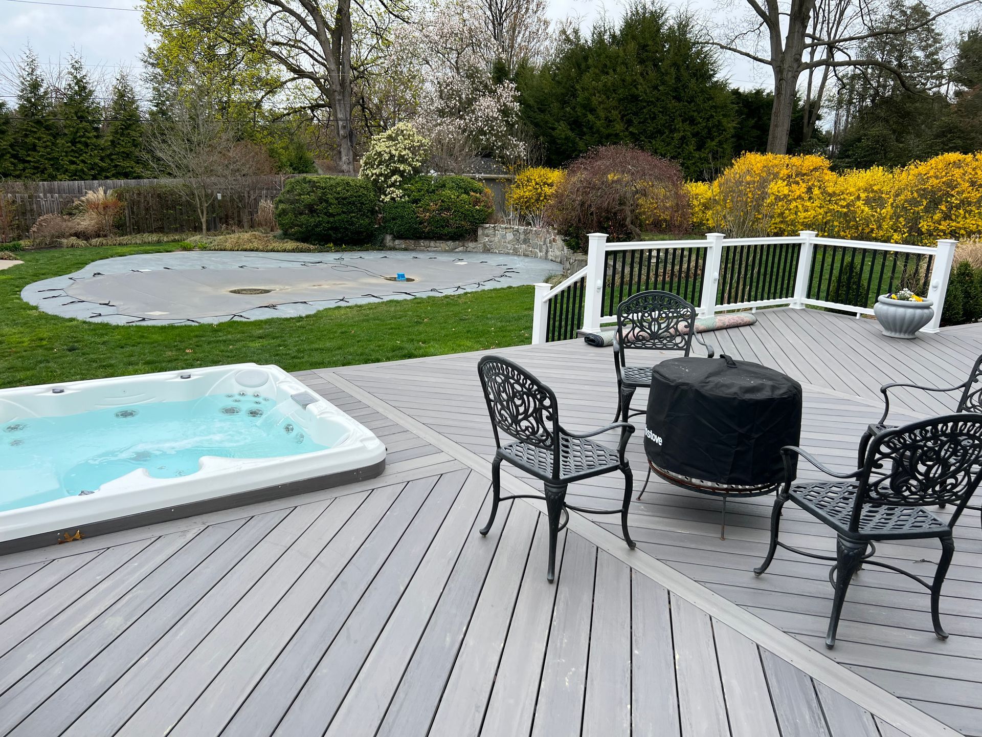 A grey composite deck with a hot tub, a fire pit, and black patio chairs, overlooking a backyard with a covered pool.