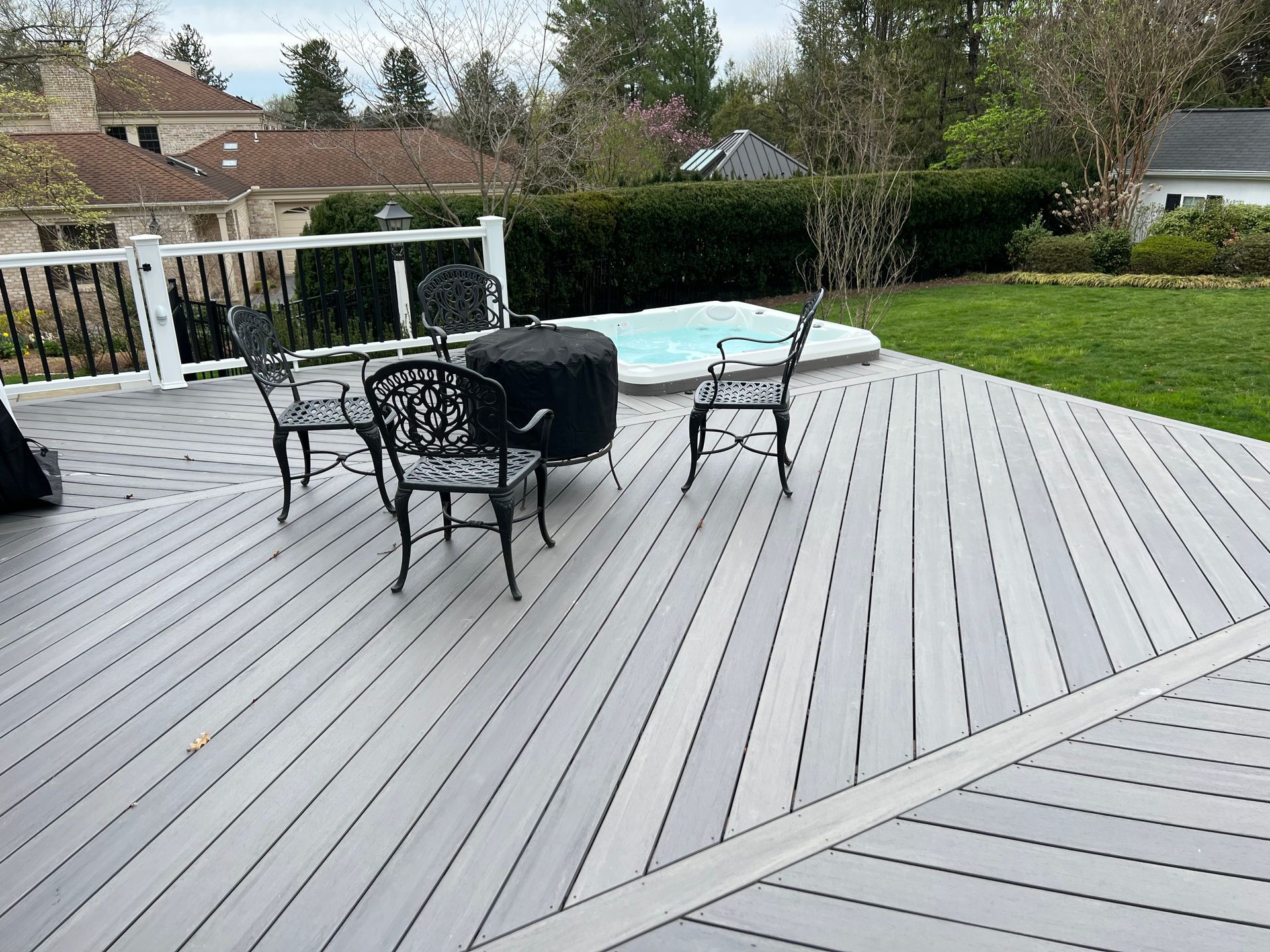 A gray deck with a small seating area, a covered fire pit, and a hot tub, overlooking a grassy backyard.