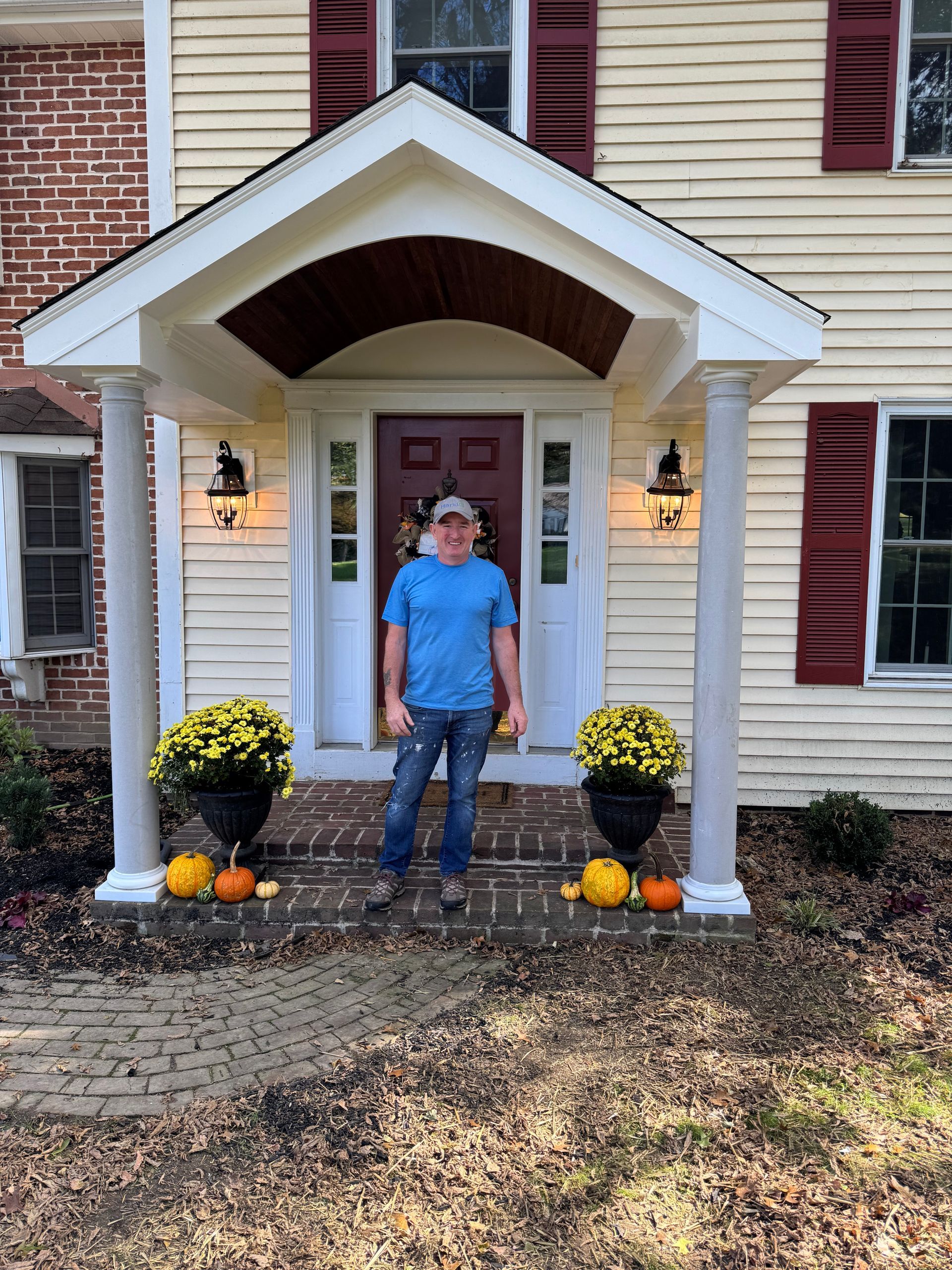 A person wearing a blue shirt and jeans stands on the brick steps of a front porch decorated with flowers and pumpkins.