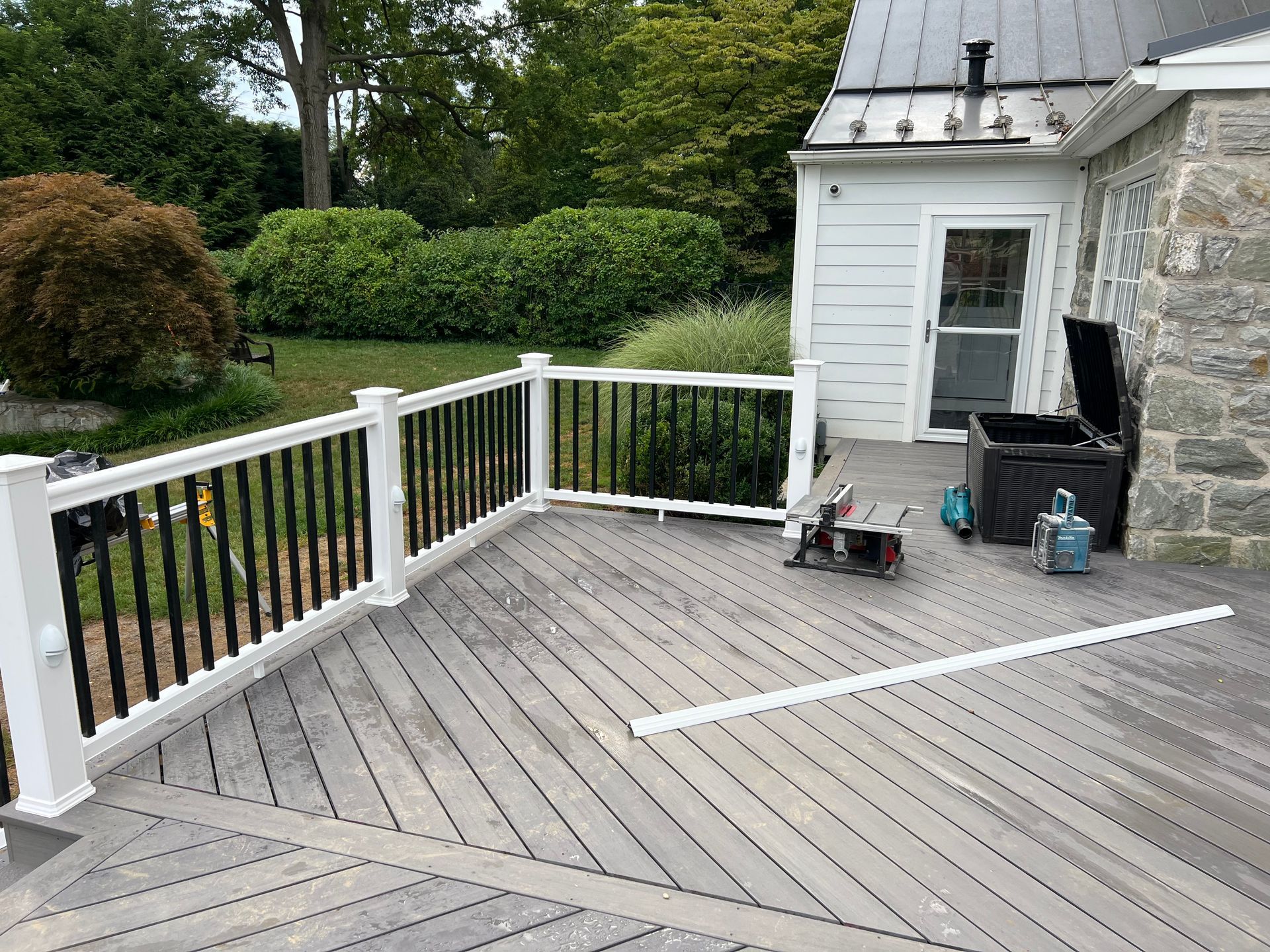 A gray wooden deck with white railings and black balusters, featuring a miter saw and a storage bin near a stone house.