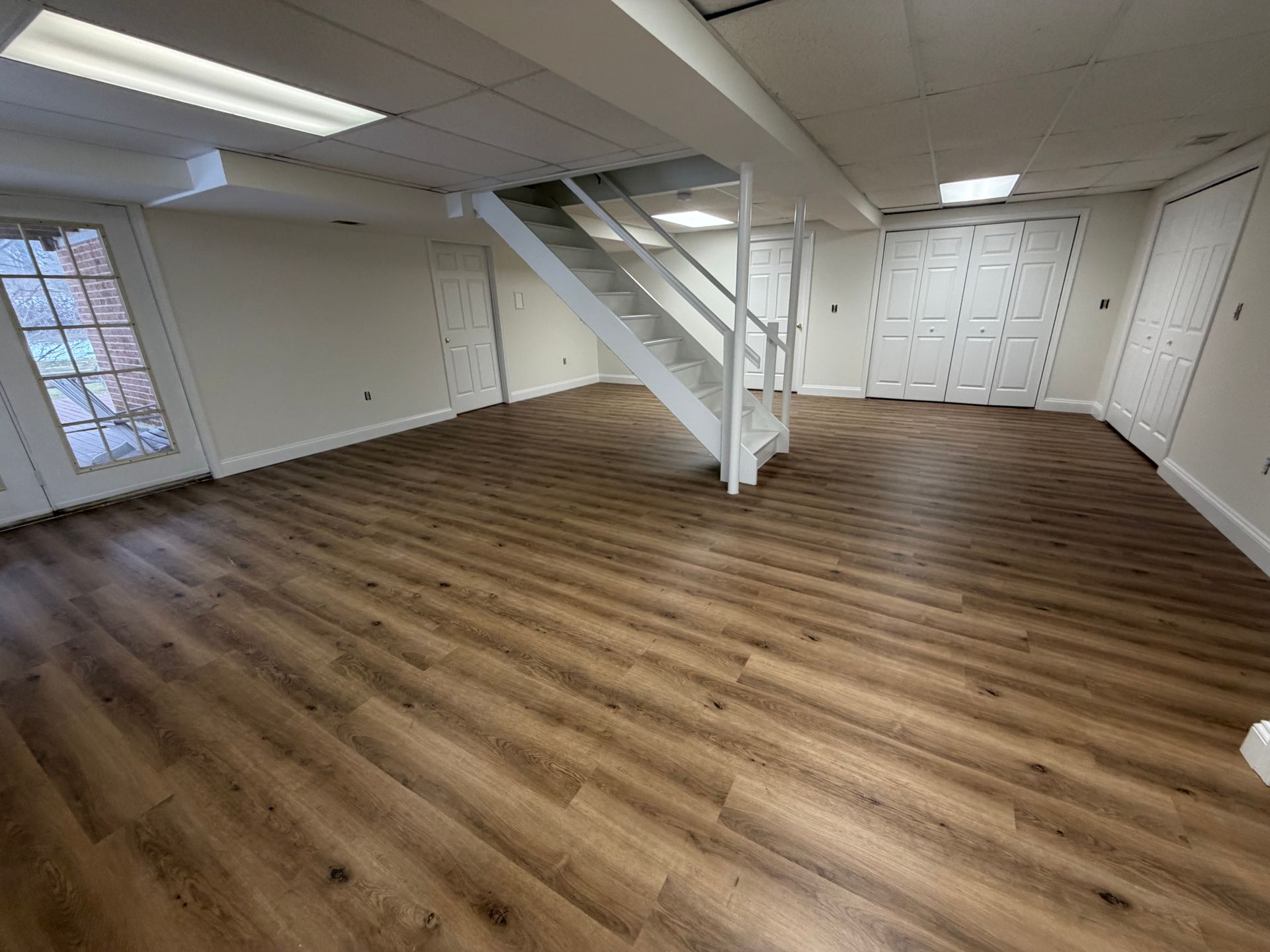 A bright basement room with wood-look flooring, white walls, recessed ceiling lights, and a white staircase.
