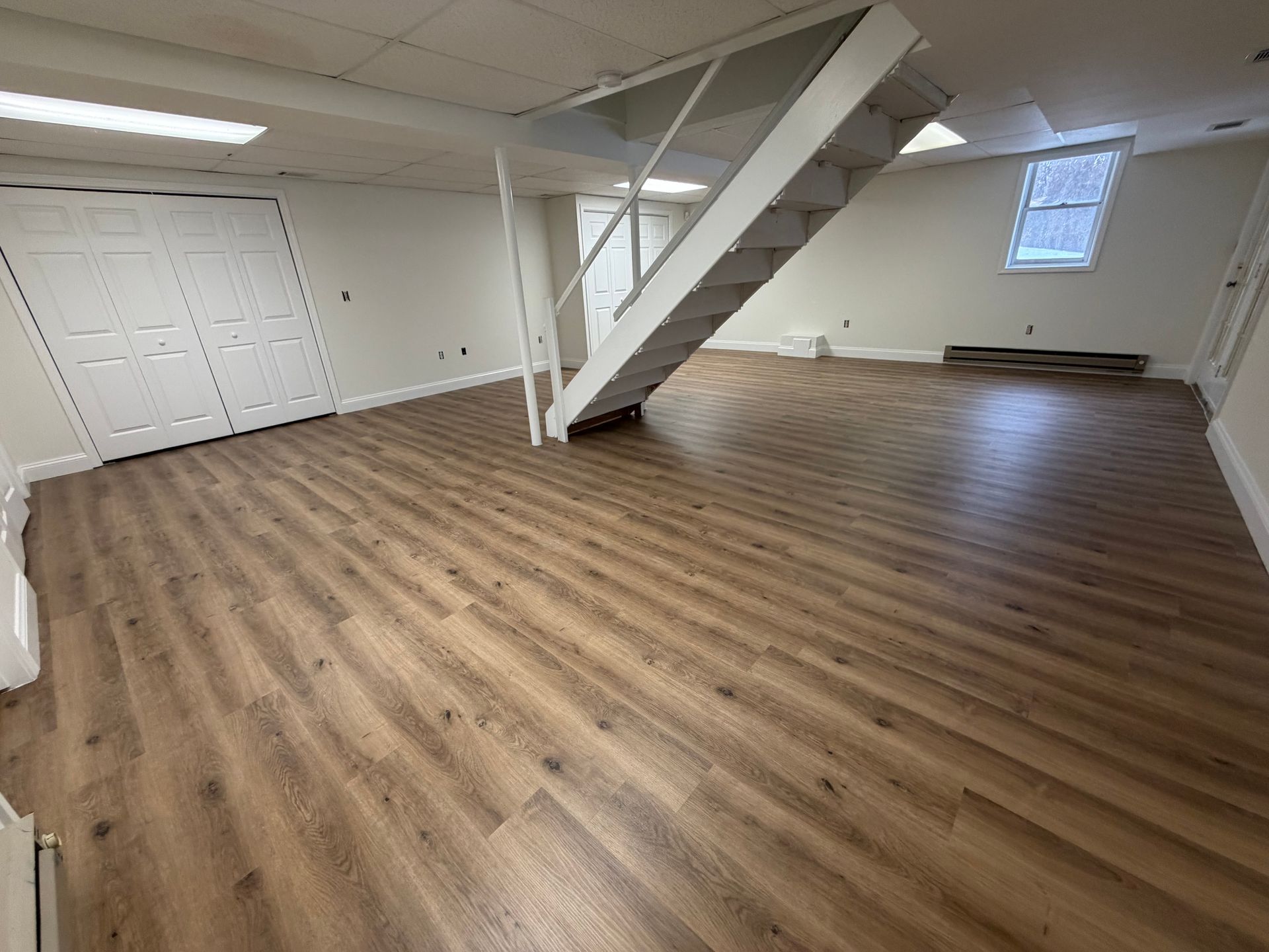 A spacious, modern basement with light-toned wood-look vinyl flooring, white walls, white closet doors, and stairs.