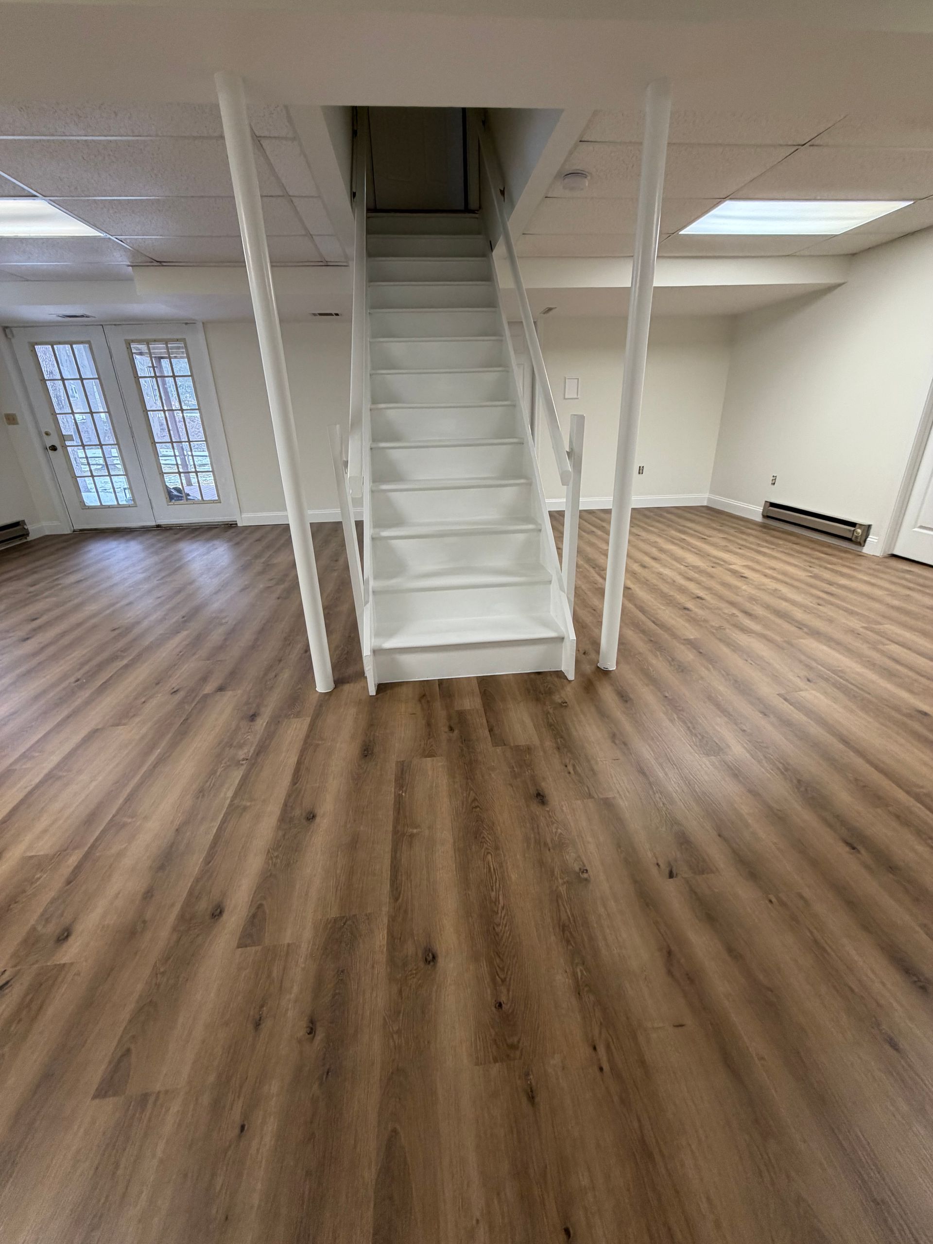 A finished basement with light wood-look flooring, white walls, recessed ceiling lights, and a white staircase.