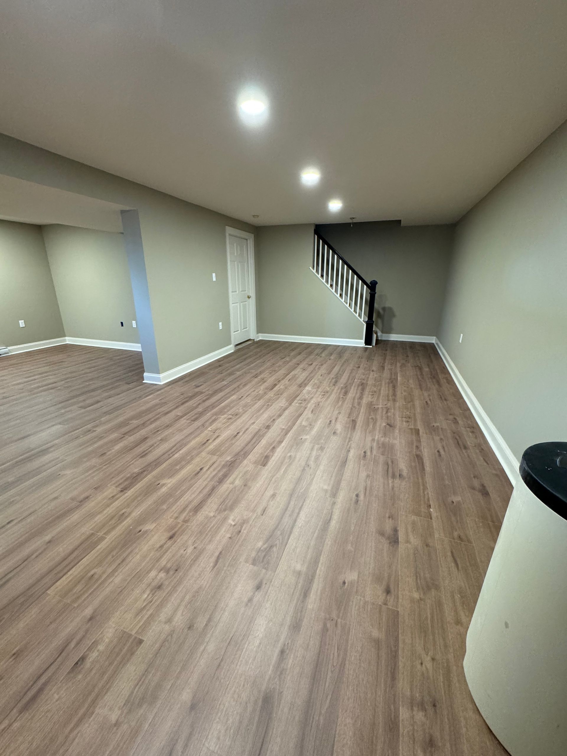 A wide view of a finished basement featuring light wood flooring, neutral walls, recessed lighting, and a staircase.