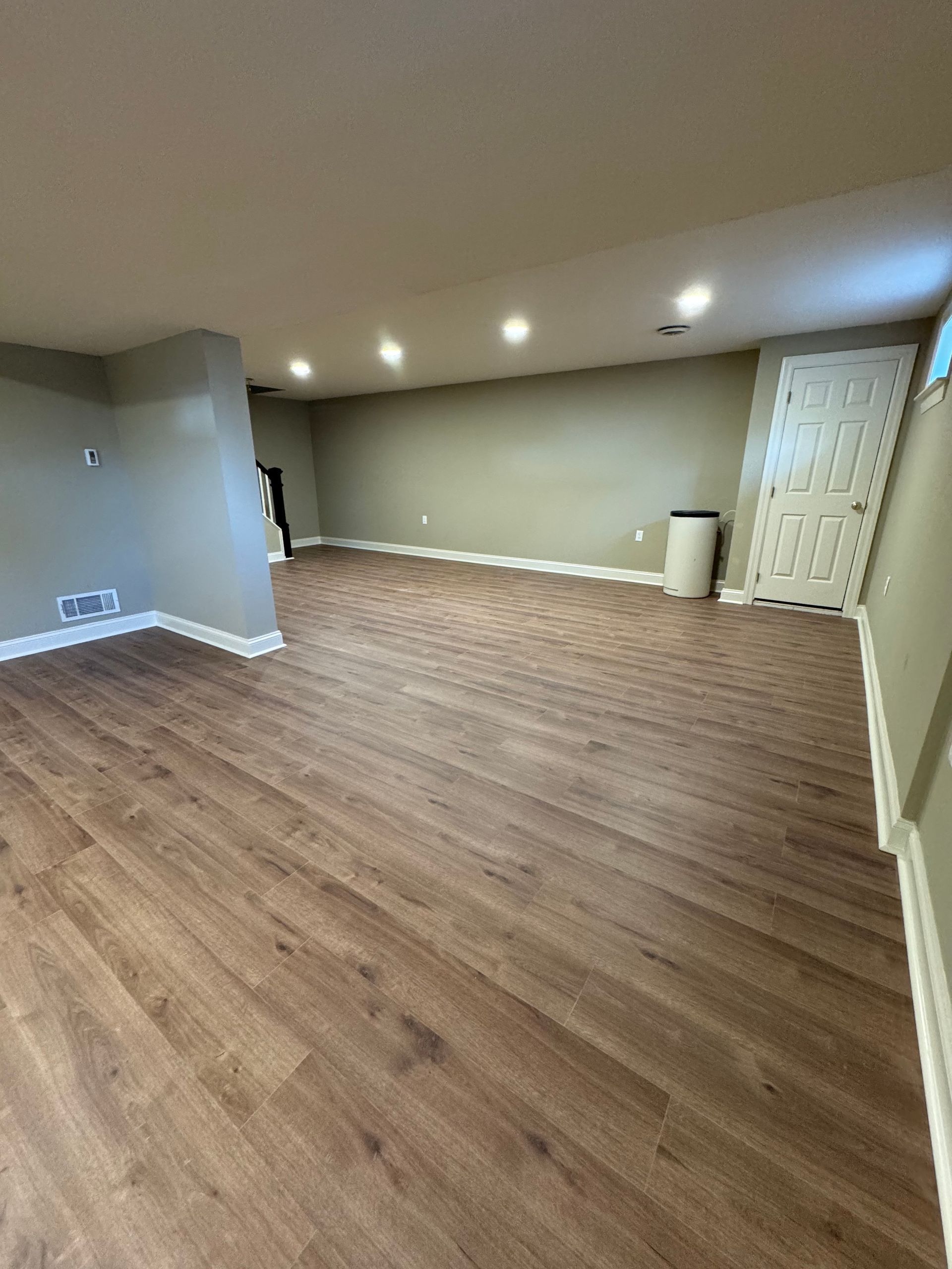 An empty basement room with light brown wood-look flooring, light grey walls, recessed ceiling lighting, and a white door.