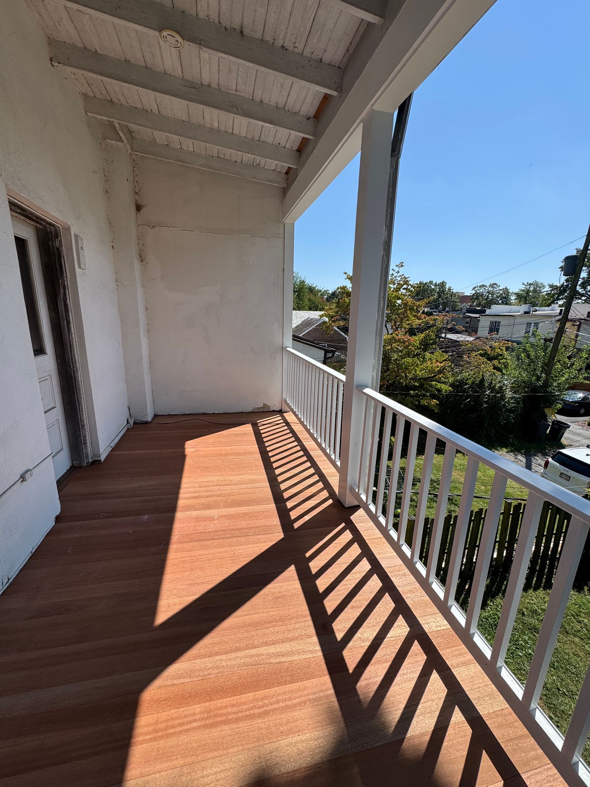 A sunlit wooden balcony with a white railing overlooks a neighborhood scene under a clear blue sky.