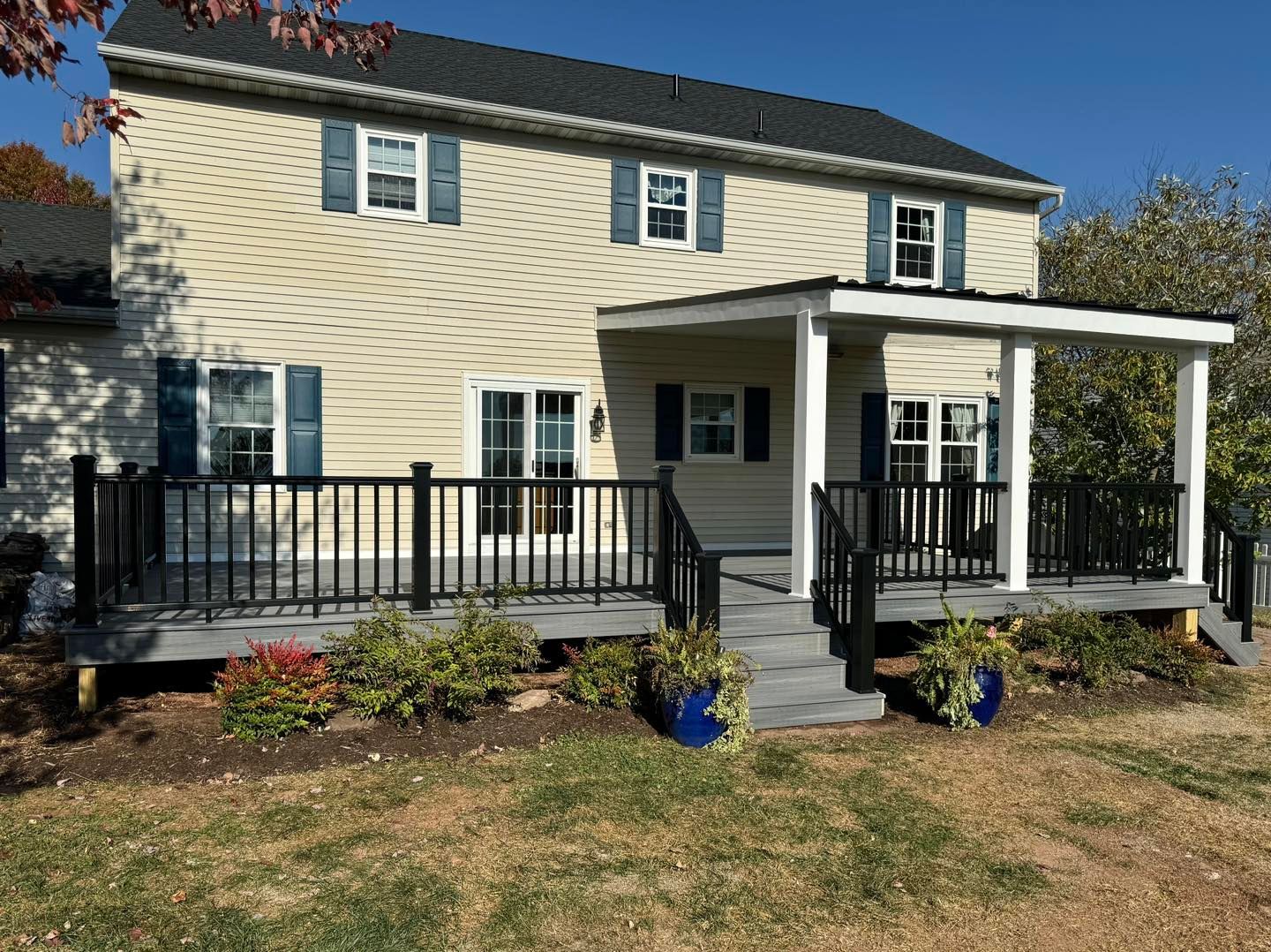 A beige two-story house with a black shingled roof, featuring a gray wooden deck with black railings and a covered porch.