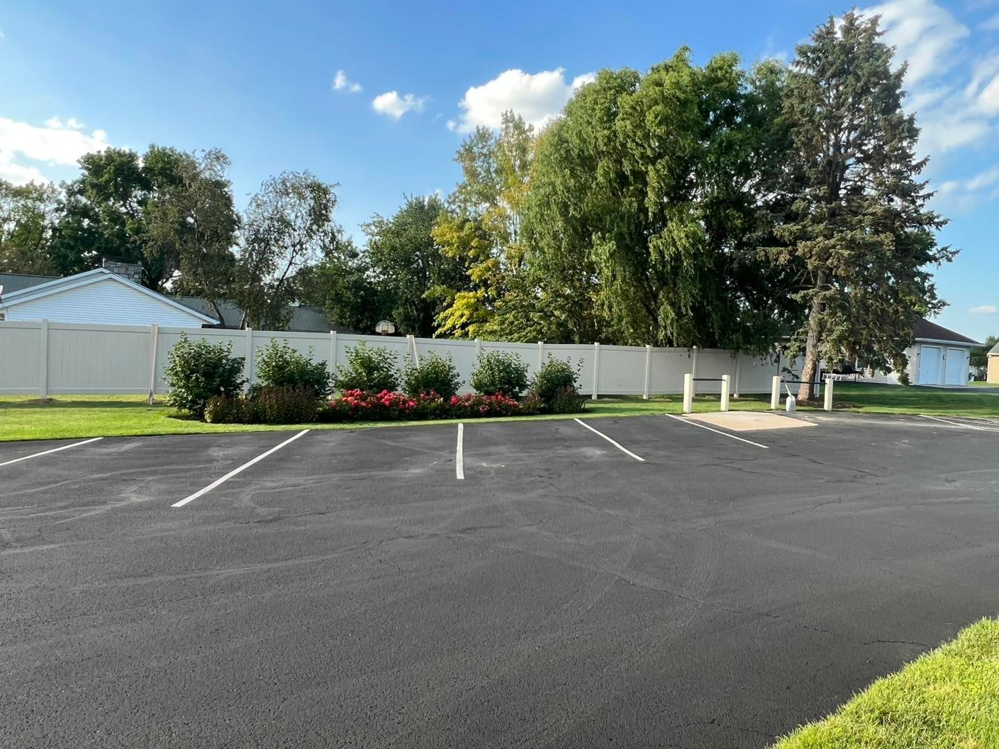 An empty asphalt parking lot with white-lined spaces in front of a white privacy fence, landscaping, and tall trees.