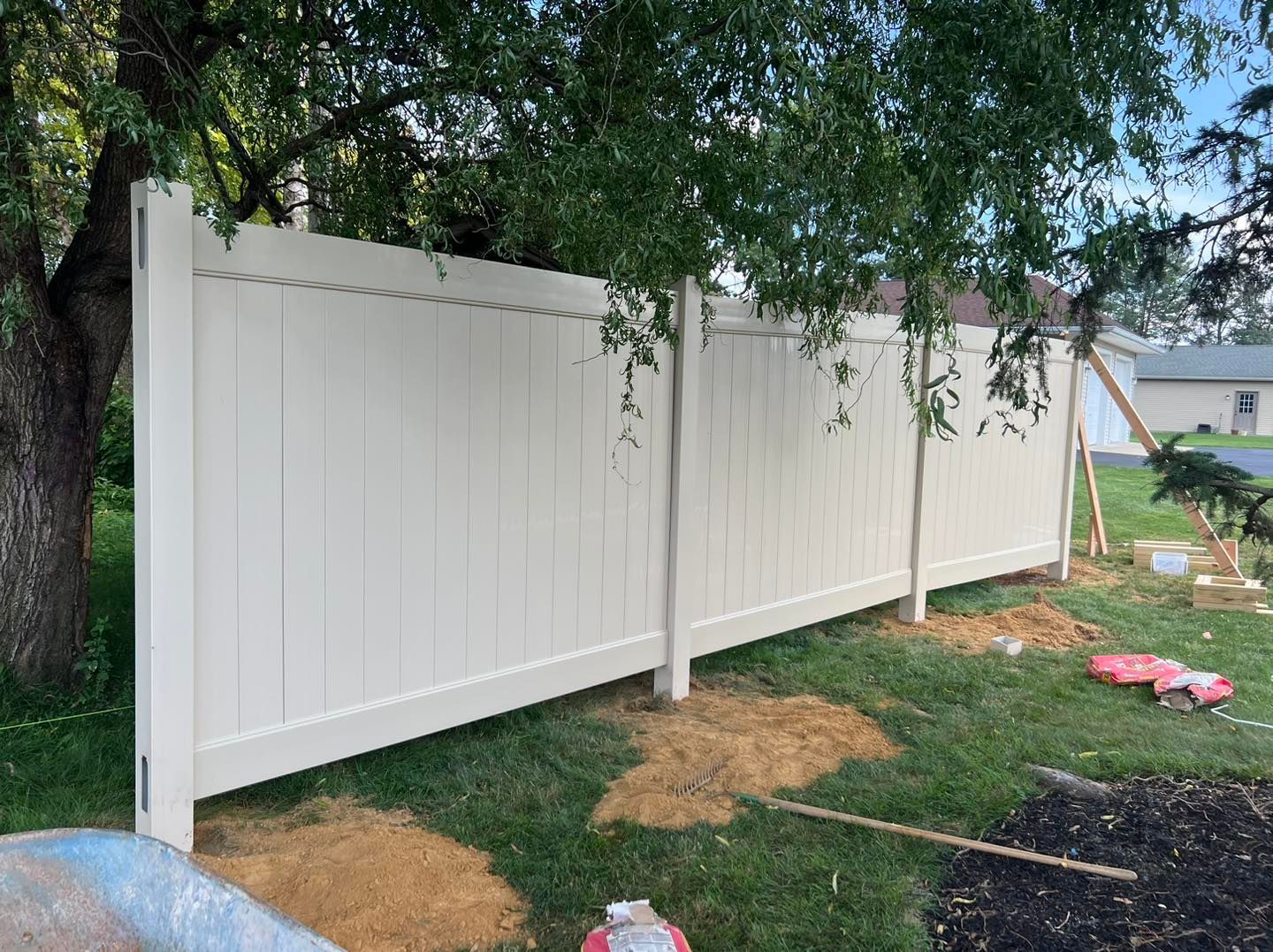 A newly installed white vinyl privacy fence stands in a grassy yard under the shade of a large tree.