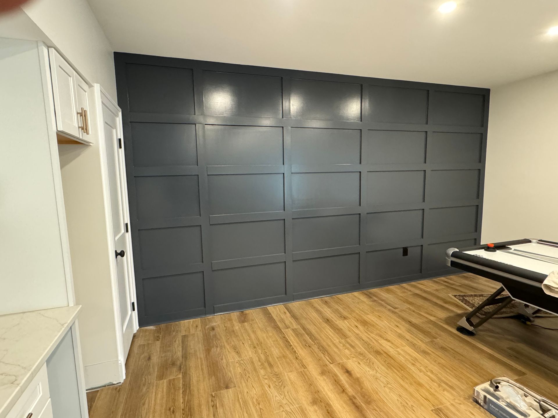 A dark gray accent wall with grid-style molding in a room with light wood flooring and a pool table visible on the right.